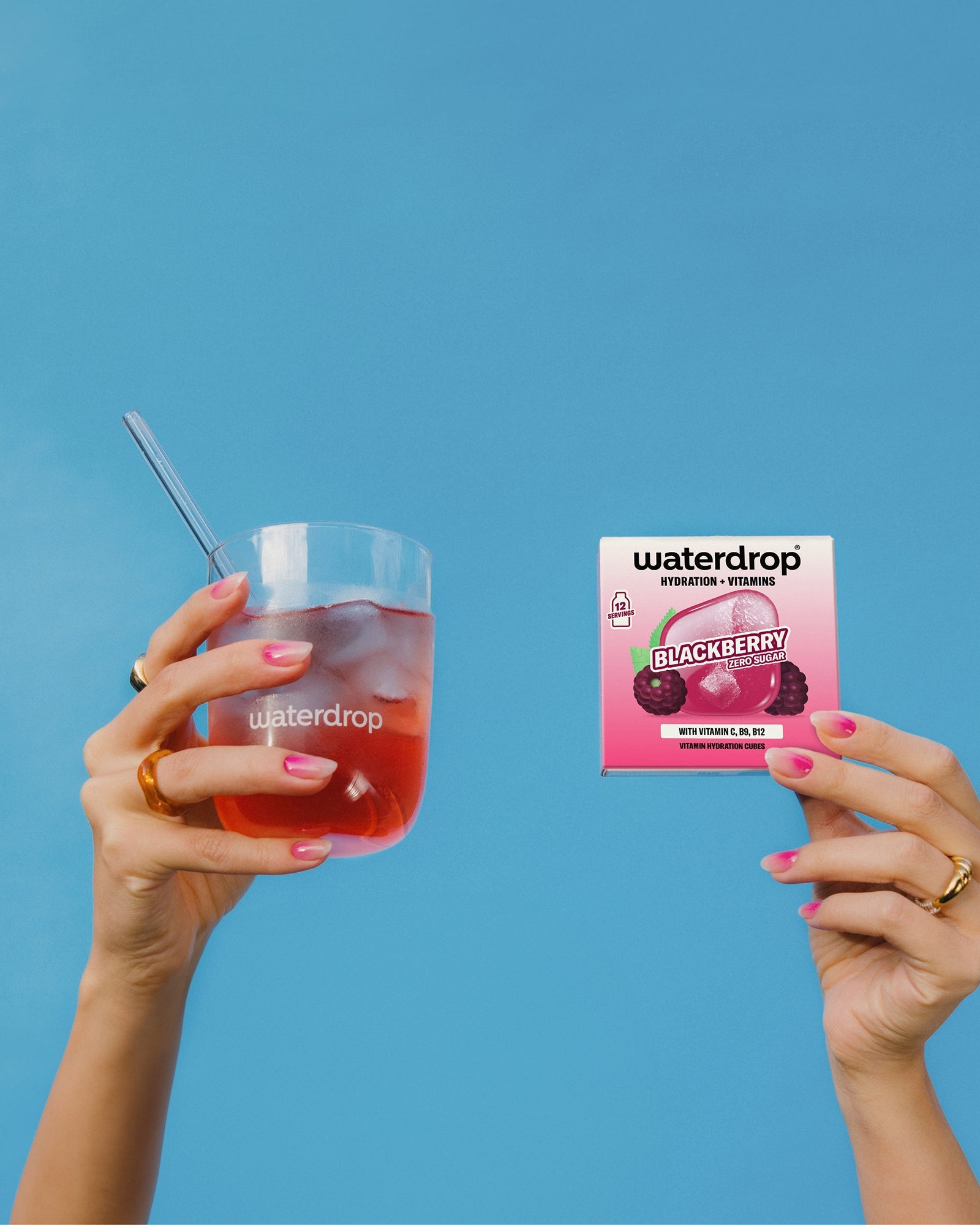Twee handen tonen de Set voor warme zomerdagen van Waterdrop®: een glas met roze drank en een doos Blackberry Zero Sugar hydratatieblokjes, ideaal voor perfecte zomerse hydratatie.