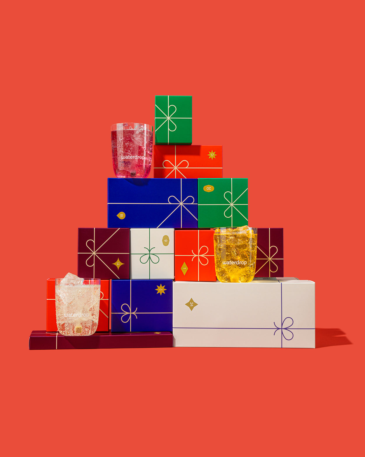 Piramide van feestelijk verpakte dozen voor de Large adventskalender, met drie gevulde Waterdrop glazen ervoor, tonen de microdrink-ervaring en uitnodigen uit tot een verfrissende, feestelijke hydratatiebeleving.
