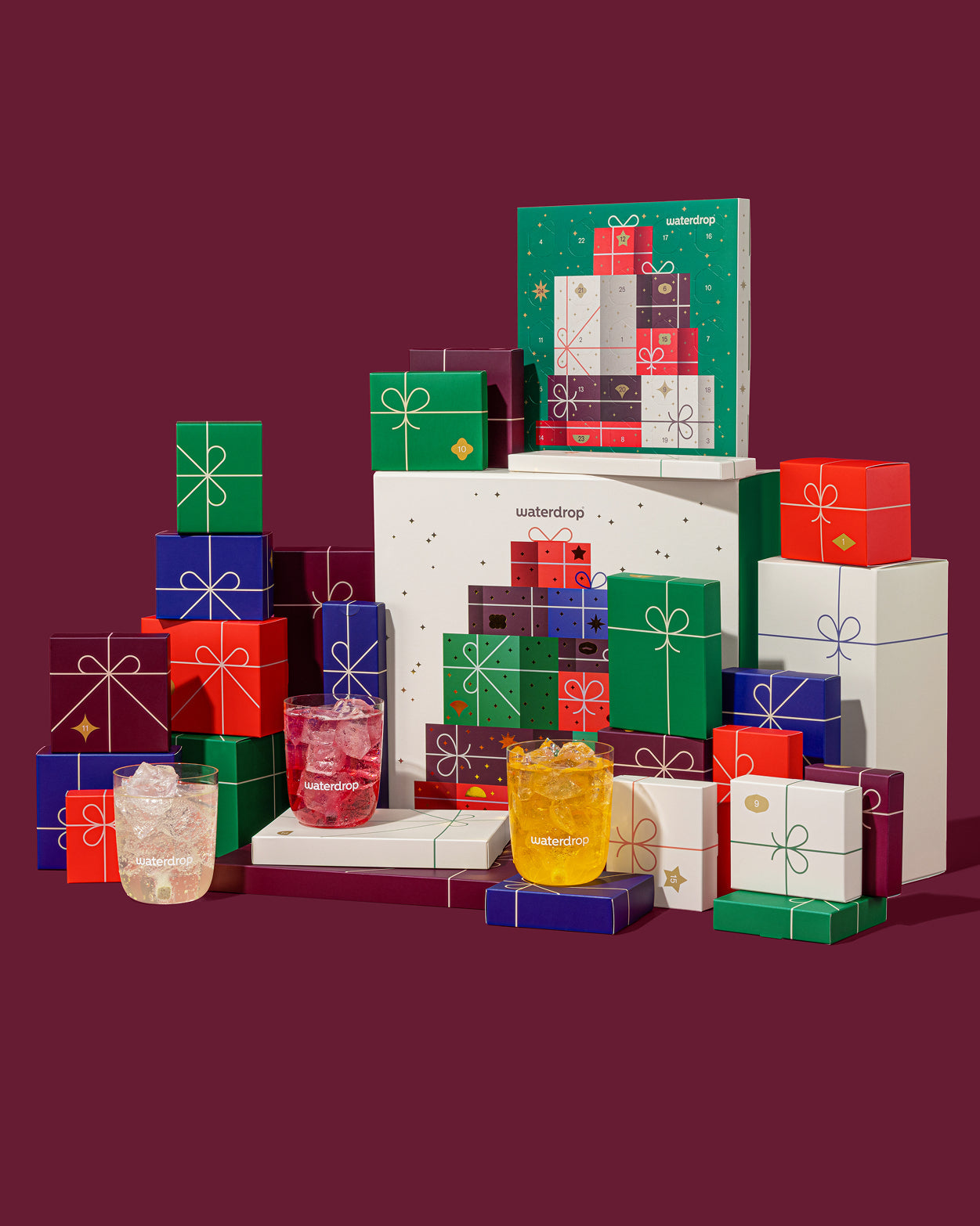 De Adventskalenderset toont een piramide van genummerde, kleurgecodeerde cadeaudoosjes en drie met ijs gevulde waterdrop-tumblers met sprankelende drankjes, passend bij de feestelijke essence van waterdrop®.