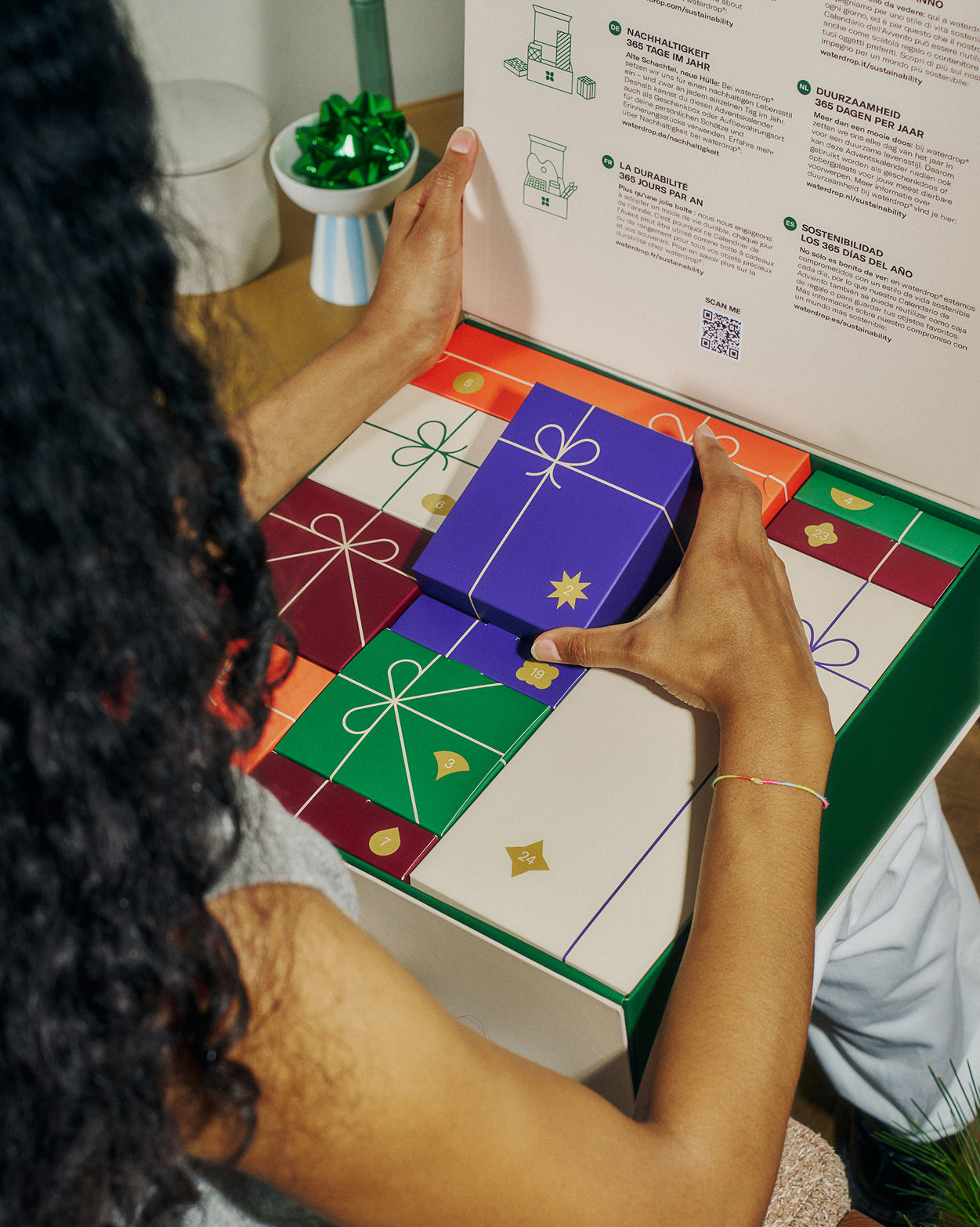 Een persoon opent de Adventskalenderset van waterdrop®, onthullend een paarse doos gemarkeerd met 19 uit een reeks van 24 luxe geschenkdoosjes. De binnenkant van het deksel benadrukt duurzaamheid.