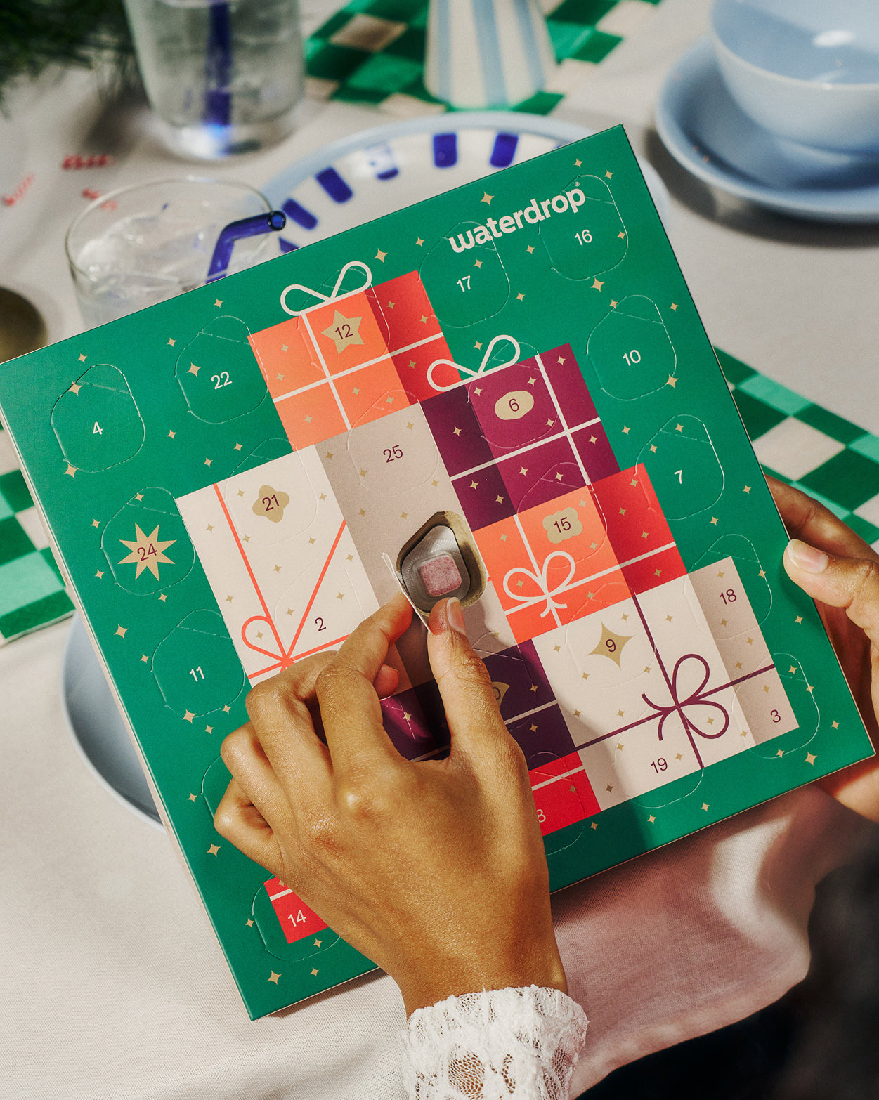 Small adventskalender van Waterdrop op tafel, met 25 genummerde vakjes. Iemand opent een vakje, onthullend een roze microdrink cube. Achtergrond toont eenvoudig tafelarrangement met glazen en servies.