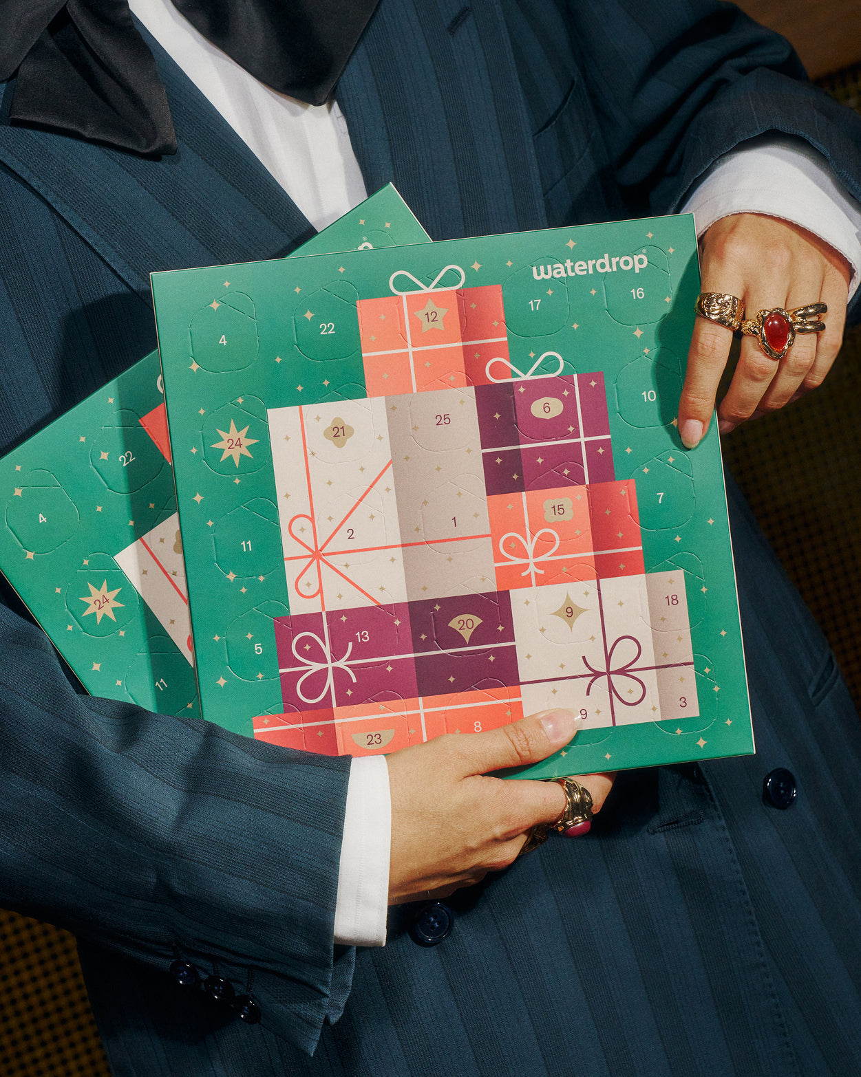 Persoon in krijtstreeppak houdt twee waterdrop® Small adventskalender duopakket vast, gevuld met 25 fruitige blokjes voor feestelijke hydratatie. Elke kalender heeft genummerde deurtjes voor dagelijks openen richting kerst.