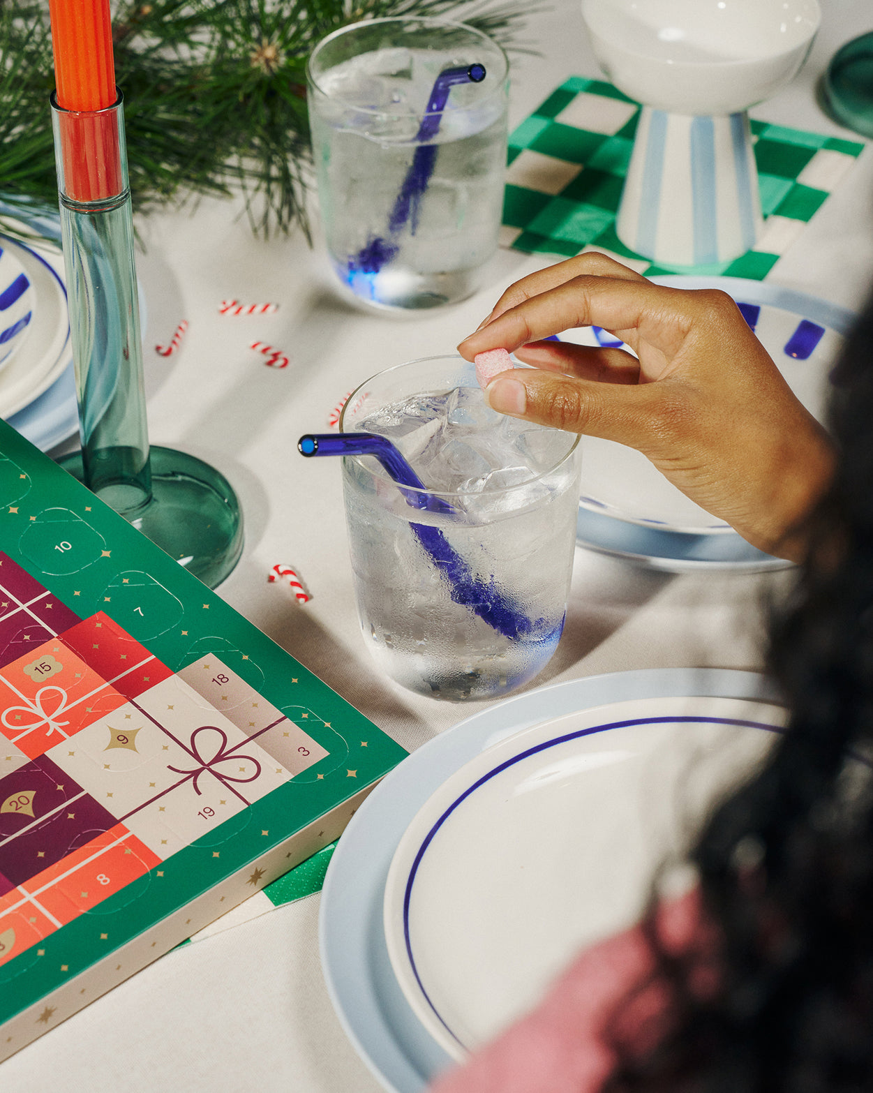 Een hand laat een roze waterblokje uit het Small adventskalender duopakket in een glas met ijswater vallen. Omringd door adventskalender, snoepjes en feestelijke tafelsetting, perfect voor gezamenlijke hydratatie.