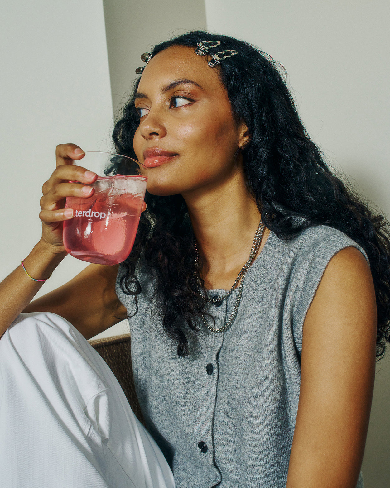 Jonge vrouw drinkt uit een waterdrop beker met helder roze drank, passend bij het Small adventskalender duopakket voor feestelijke fruitige hydratatie, terwijl ze een grijze top en witte broek draagt.