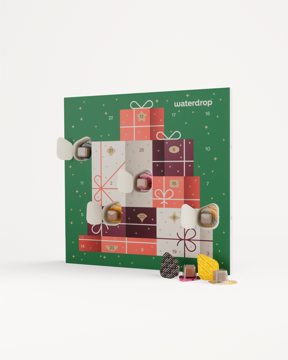 Kleine adventskalender van waterdrop® met 25 vensters in cadeaupatroon. Enkele vensters geopend, onthullend folie-verpakte microdrinkblokjes. Voor de kalender liggen los verpakte smaakblokjes, ideaal voor een fruitige kerstervaring.