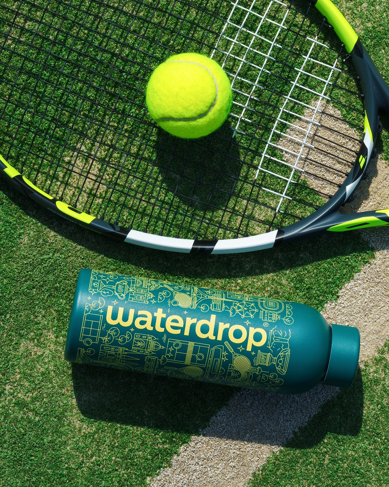 AO26 Melbourne RVS Drinkfles op grasveld naast tennisracket en gele tennisbal, met de tekst waterdrop zichtbaar. Dubbelwandig roestvrij staal, houdt dranken 24 uur koud of 12 uur warm.