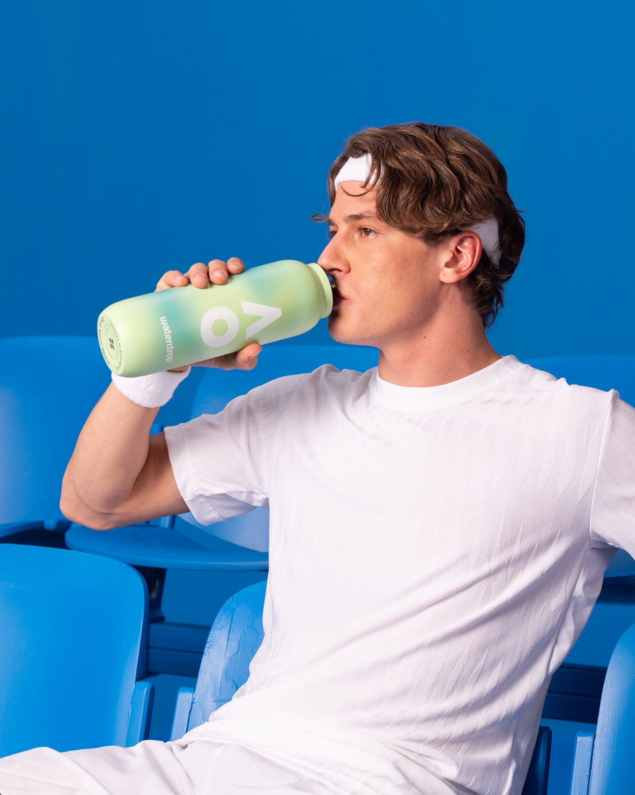 Jonge man in witte sportkleding drinkt uit de AO26 Performance All-Purpose Thermo-fles, zittend op blauwe stoelen. Deze duurzame fles van waterdrop® houdt dranken tot 24 uur ijskoud.