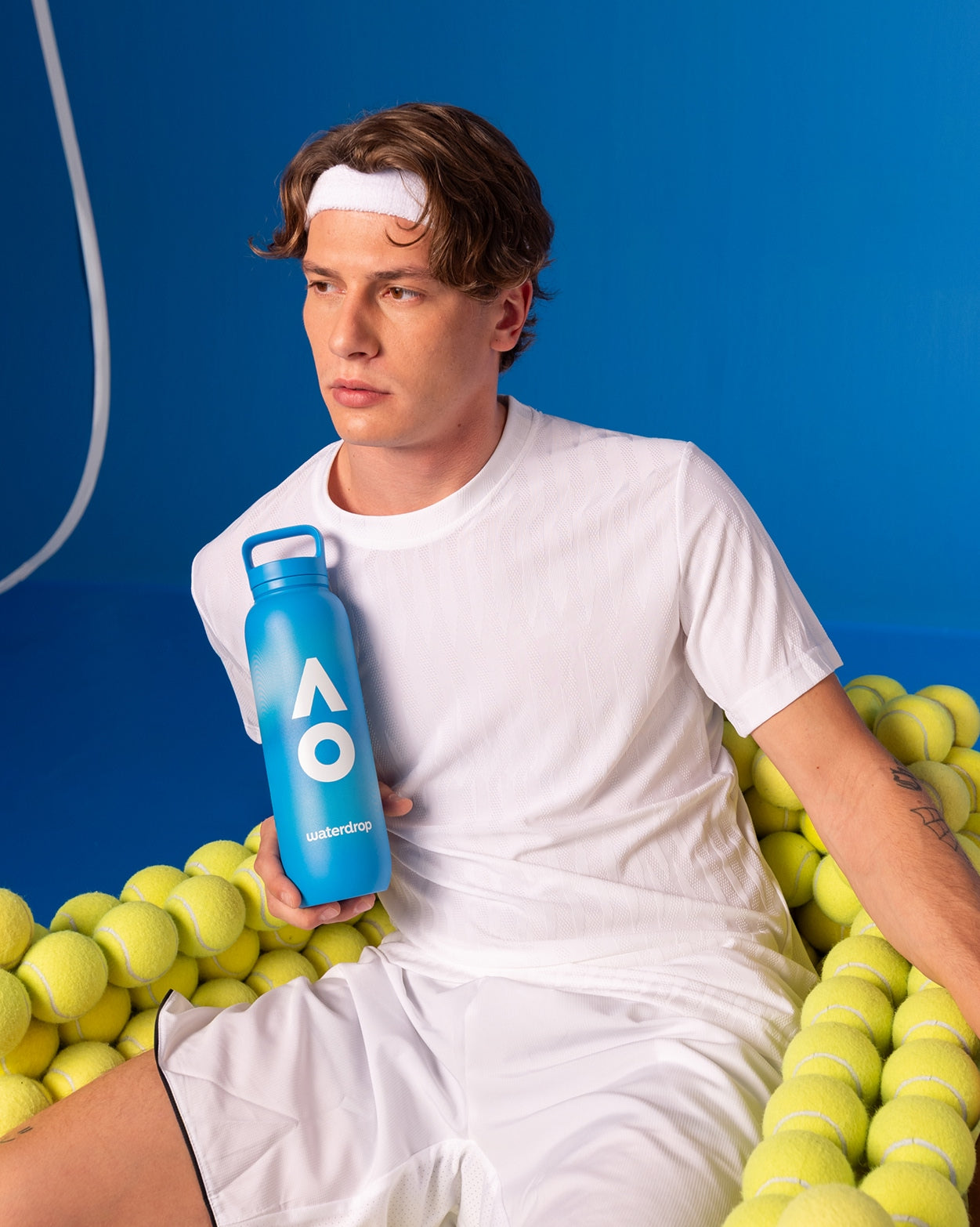 Jonge man in witte sportkleding zit in tennisballen, houdt AO26 Officiële Spelersfles omhoog. Deze waterdrop® fles biedt langdurige hydratatie, ideaal voor sporters.