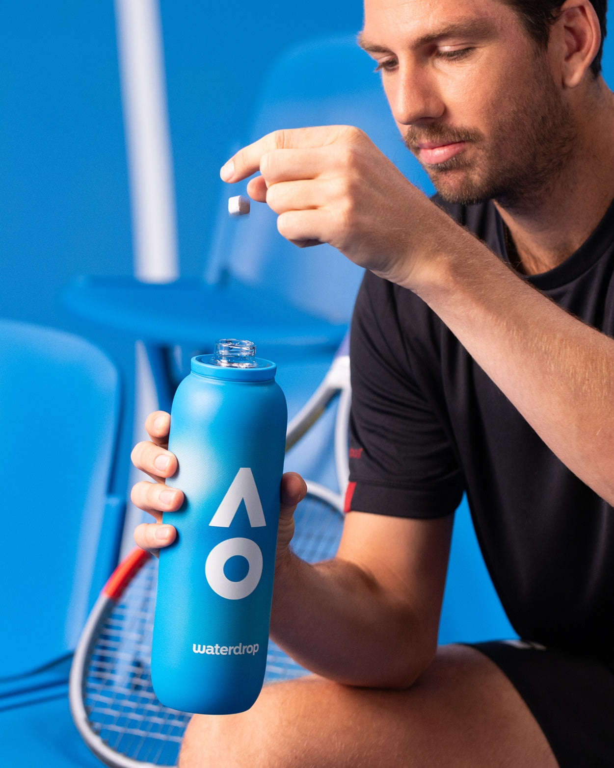 Man in tennisoutfit houdt de AO26 Officiële Spelersfles vast, klaar om een waterdrop®-blokje toe te voegen. Deze Limited Edition fles houdt drankjes 24 uur ijskoud.