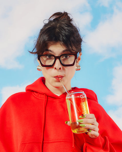 Jongere met oversized bril drinkt uit een heldere beker met herbruikbaar rietje, genietend van de Set: 3+1 Microdrinks. Nagels met lieveheersbeestjesmotief benadrukken de stijlvolle presentatie van de fruitige waterdrop®-ervaring.