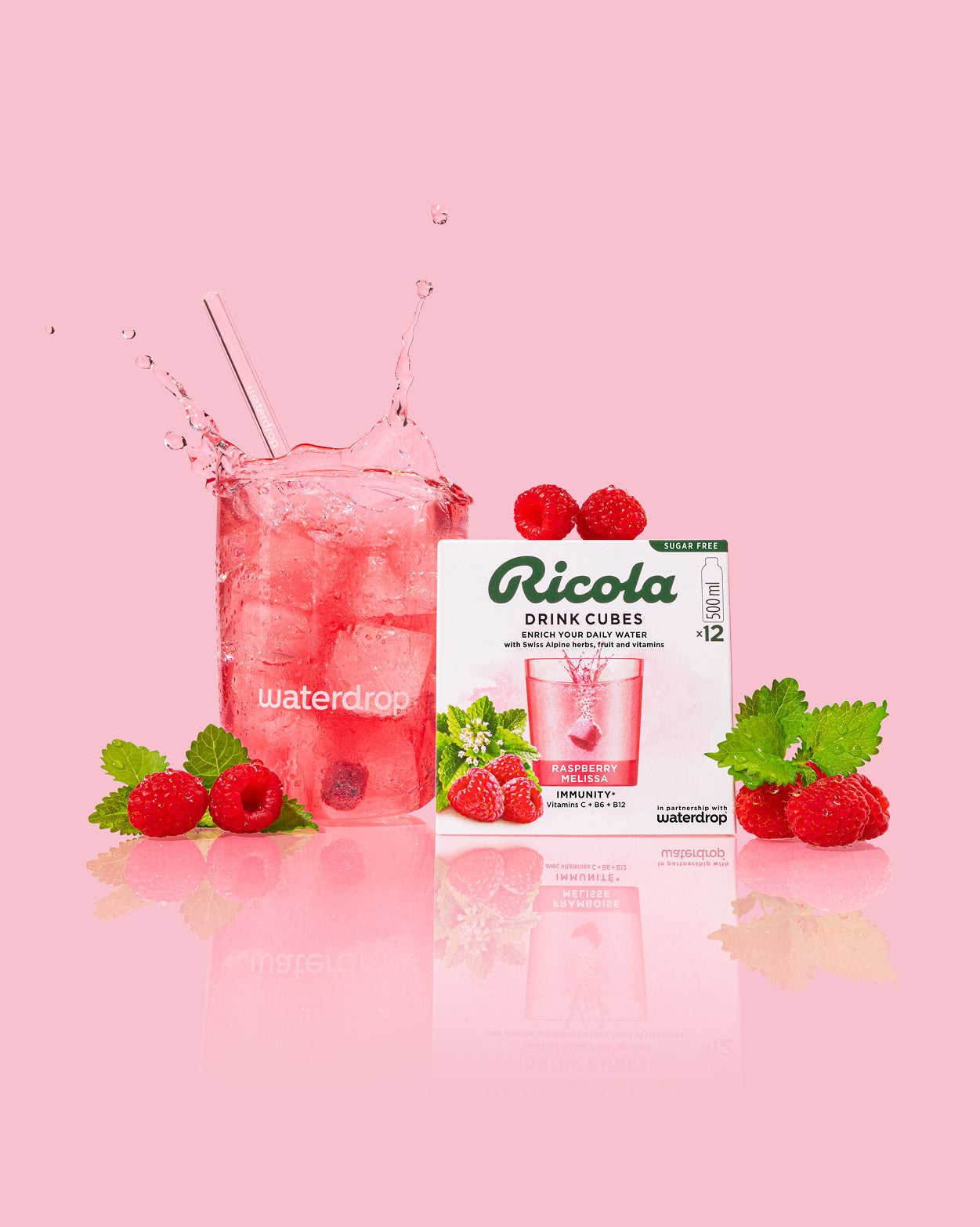 Cadeauset met Ricola en glazen fles: Een waterdrop® glas gevuld met een roze drank, omringd door Ricola Drink Cubes in Raspberry Melissa smaak, met verse frambozen en citroenmelisse.