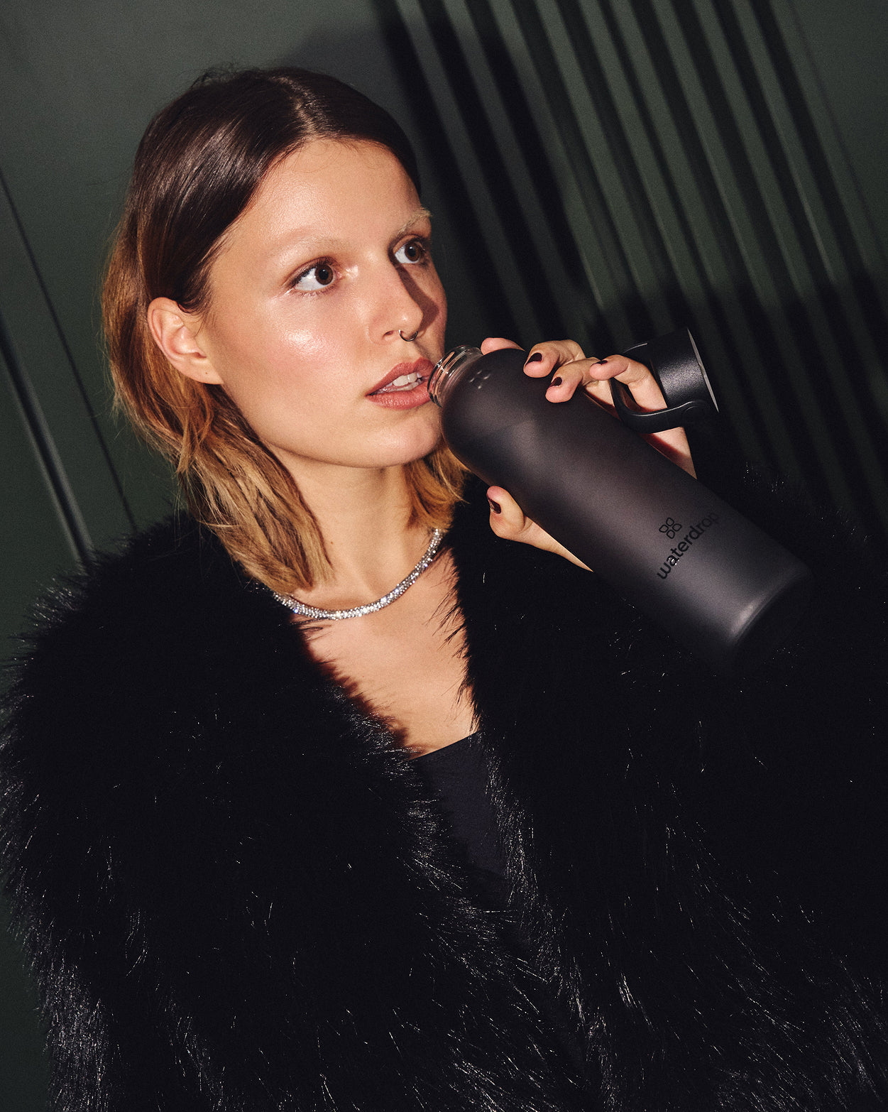 Jonge vrouw met een zwarte faux-fur jas drinkt uit een waterdrop® matte zwarte geïsoleerde fles, deel van de Cadeauset met matglazen fles, voor een stijlvolle hydratatie-ervaring.
