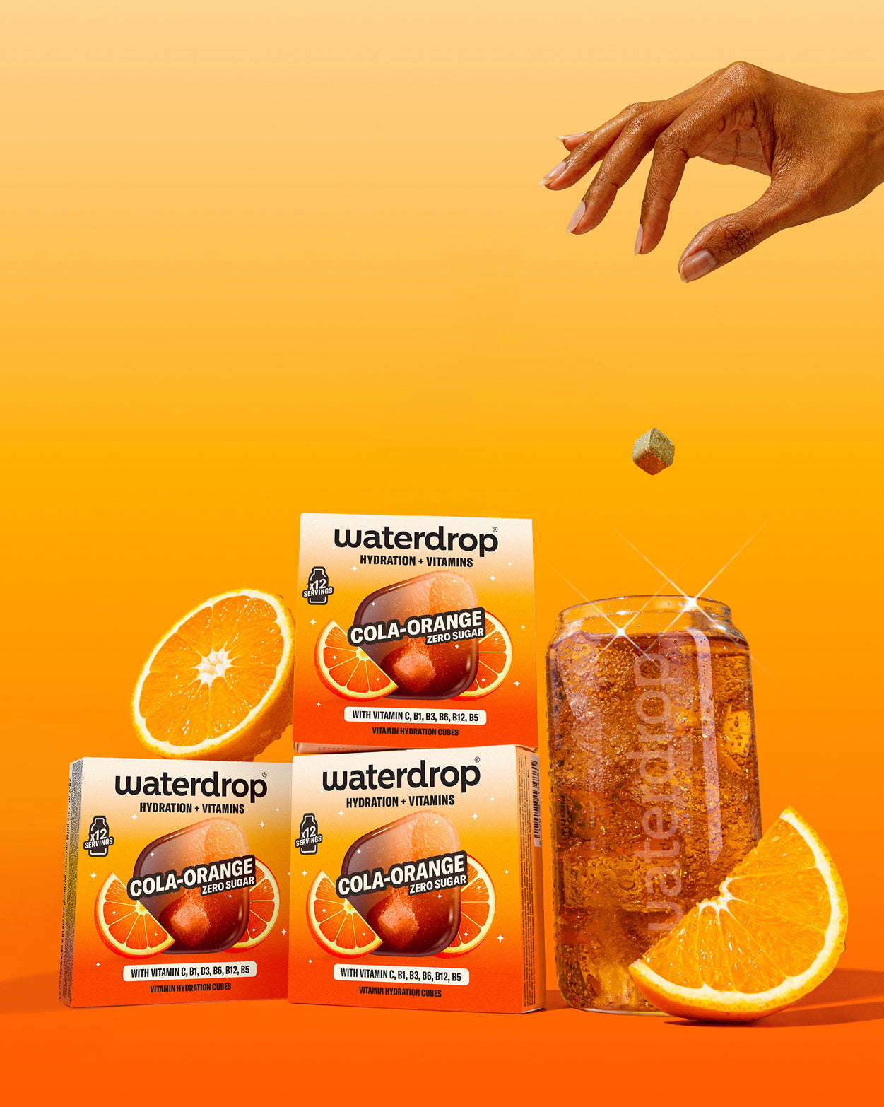 COLA ORANGE 3+1: Drie waterdrop® dozen met 12 suikervrije hydratatie- en vitamineblokjes. Een hand laat een blokje in een cola-oranje drank vallen. Verse sinaasappelschijfjes omlijsten het tafereel.