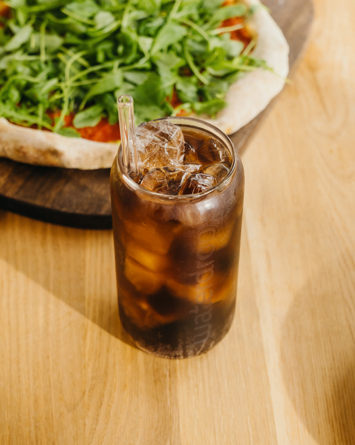 COLA Glass Can van hoogwaardig borosilicaatglas naast een glas ijsthee met rietje op tafel, biedt een duurzame, prikkelende drinkervaring. Limited Edition, ideaal voor waterdrop® NL's verfrissende assortiment.