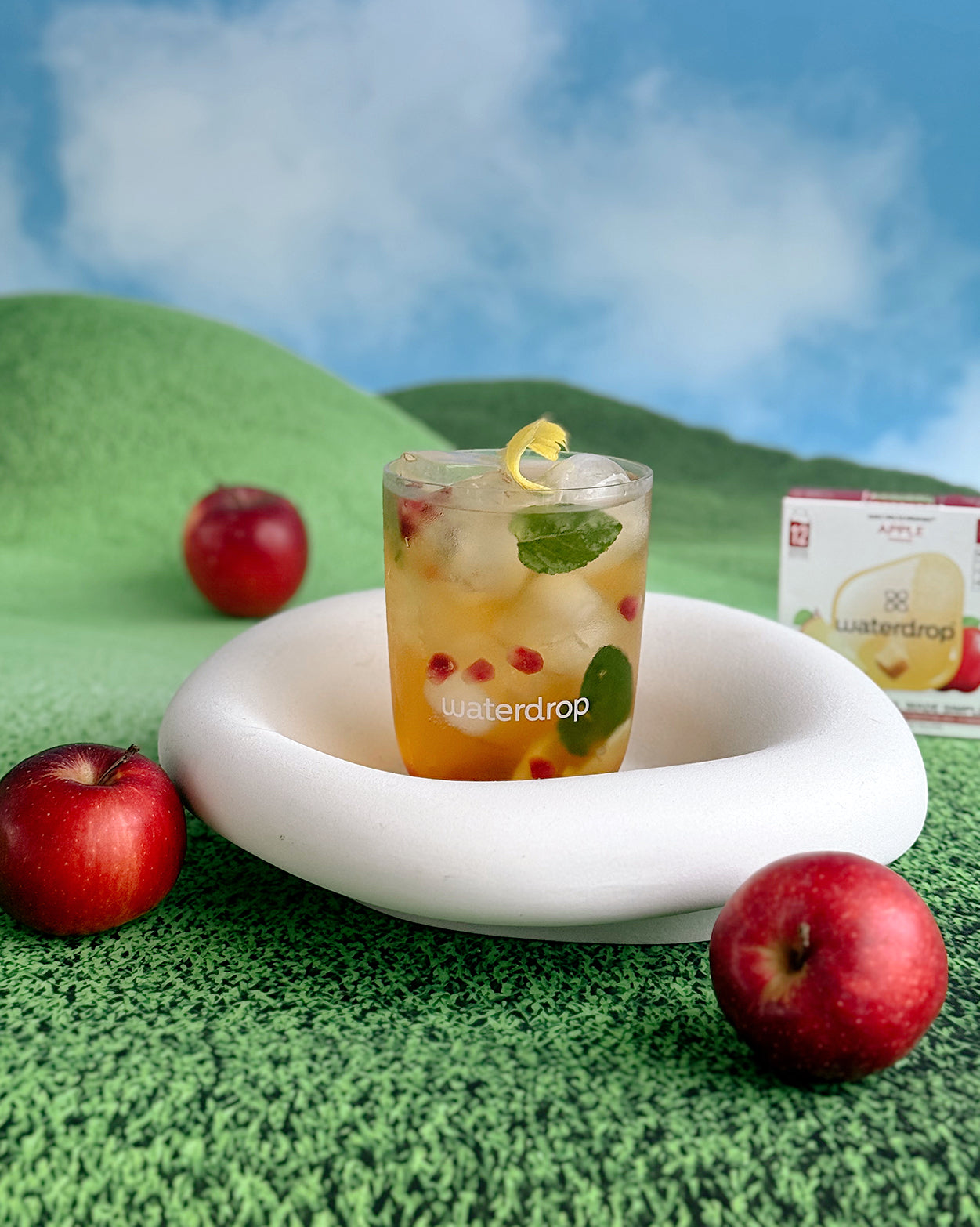 Apple Sparkle Set: Een glas met ijs, granaatappelpitjes, muntblaadjes en citroenschil op een witte standaard, omringd door rode appels en een waterdrop® appelverpakking, voor een verfrissende ervaring.