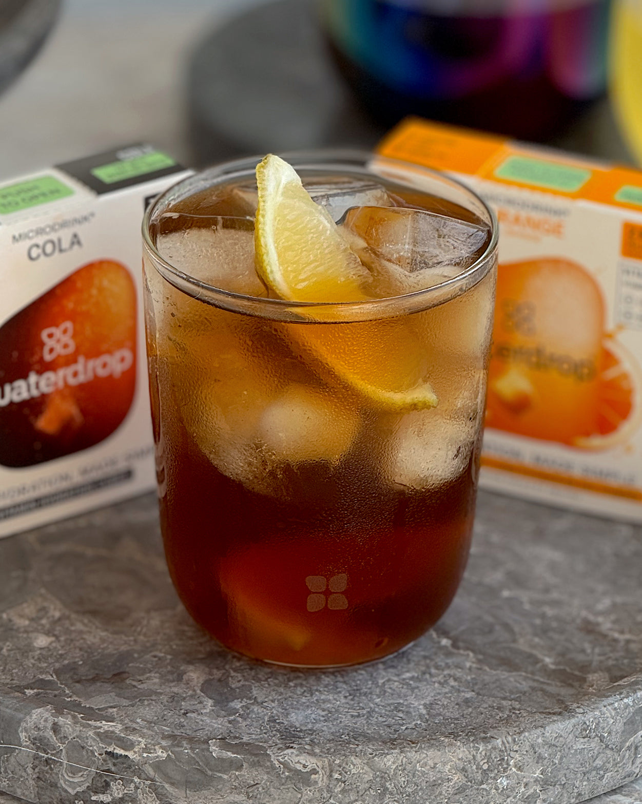 Een helder glas gevuld met een donkere, cola-gekleurde ijskoude drank, gegarneerd met een citroen- of limoenpartje, gemaakt met de Cola Orange Set 3+1 van waterdrop®, zichtbaar zijn de Microdrink-dozen Cola en Orange.