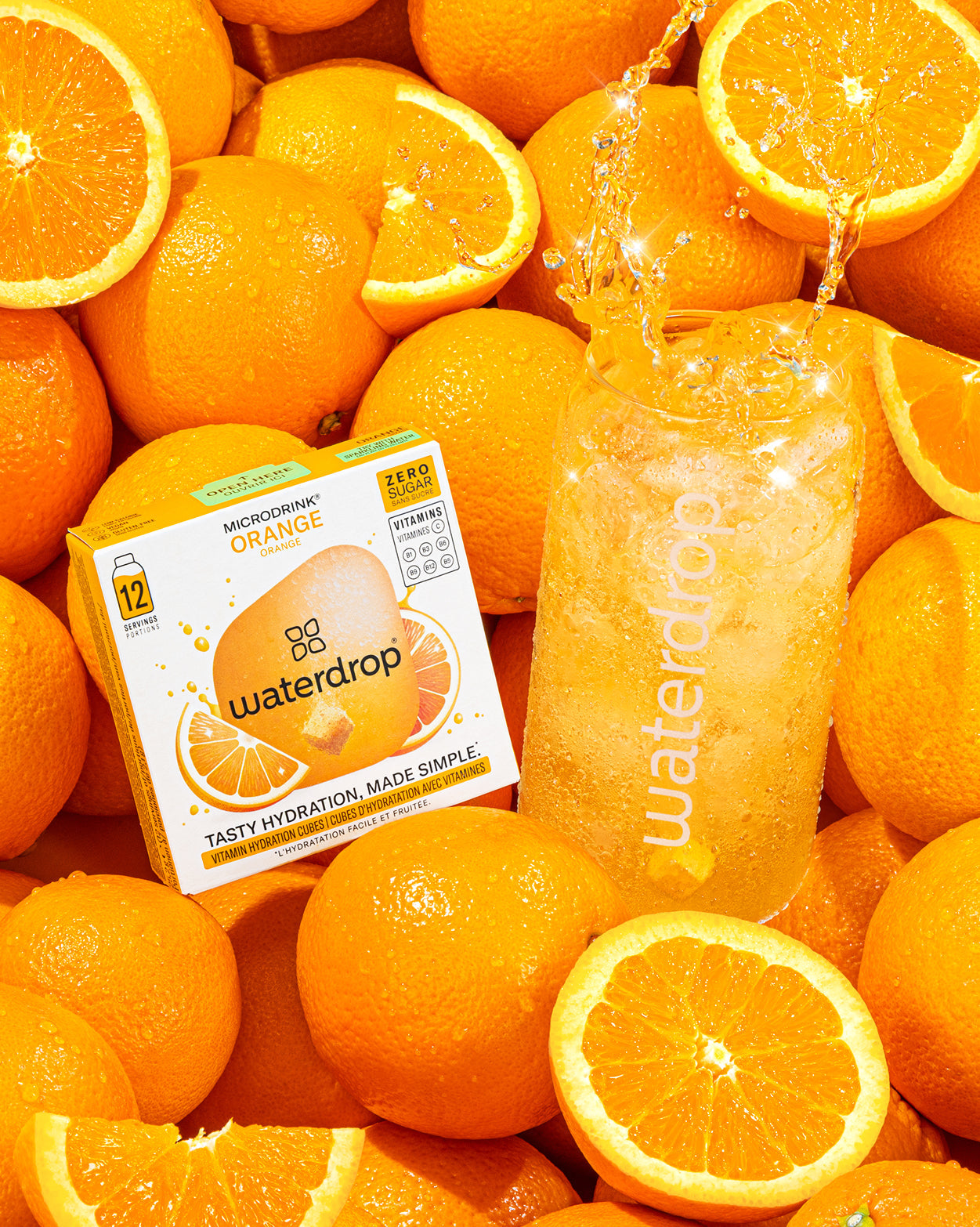 Een stapel verse sinaasappels en een Waterdrop Microdrink Orange doos naast een Waterdrop-glas met een bruisende, oranje hydratatiecube. Geniet van deze Cola Orange Set 3+1 voor verfrissende vitaminen.