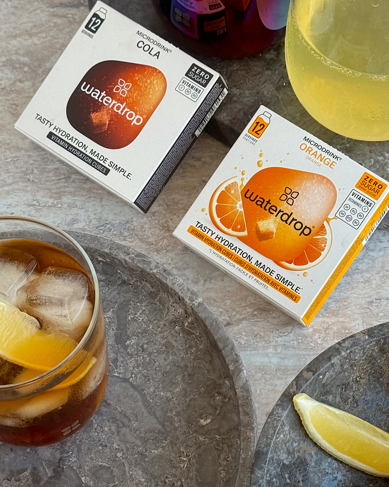 Cola Orange Set 3+1: Twee dozen Waterdrop Microdrink met twaalf suikervrije hydratieblokjes per smaak, Cola en Orange. Twee glazen drankjes met citrusschil en citroenpartje ernaast. Geniet afzonderlijk of samen.