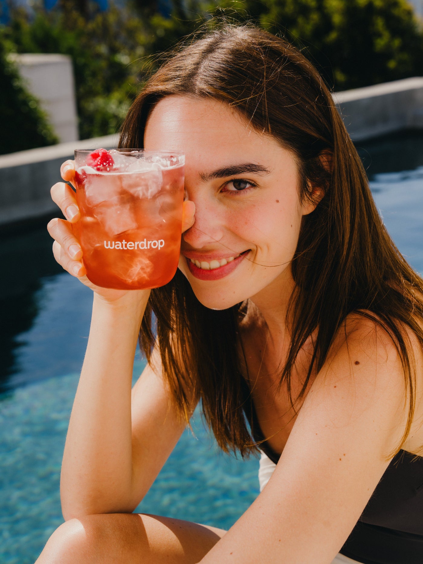 Jonge vrouw bij zwembad houdt een glas met waterdrop-logo vast, gevuld met een roze drank uit de ICE TEA Set 3-Pack, versierd met een framboos. Ze glimlacht stralend.