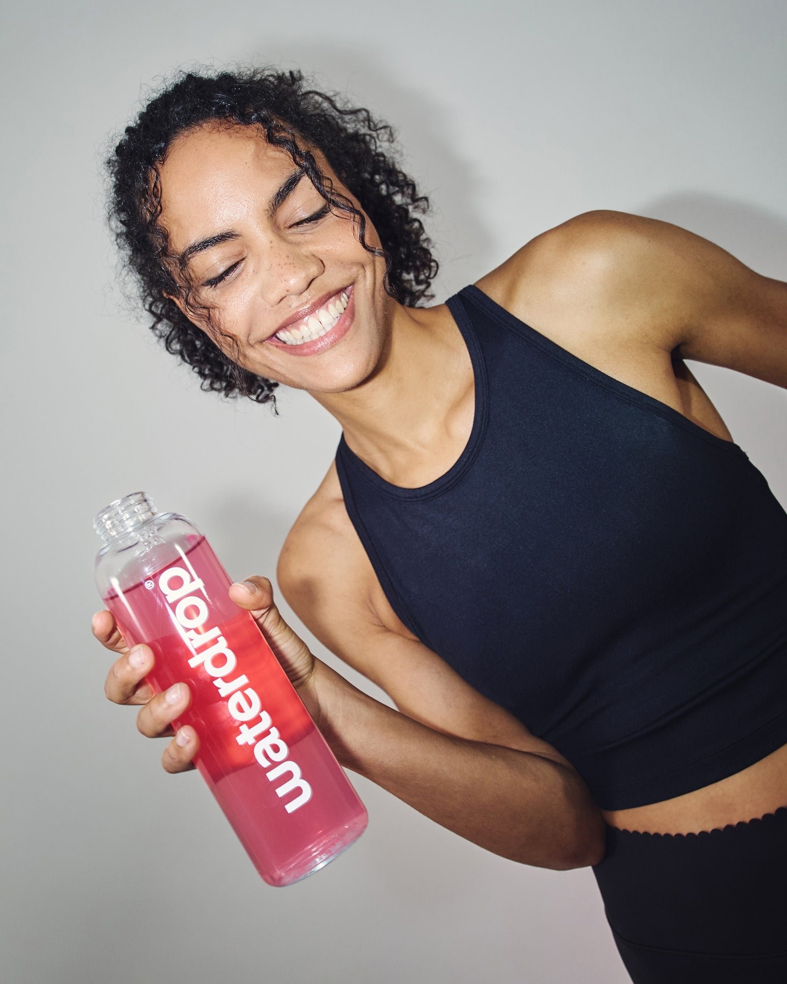 Jonge vrouw in zwarte sporttop houdt een fles met roze vloeistof omhoog, genietend van de Microlyte RECHARGE Set 3+1 met elektrolyten en vitamines van waterdrop®.