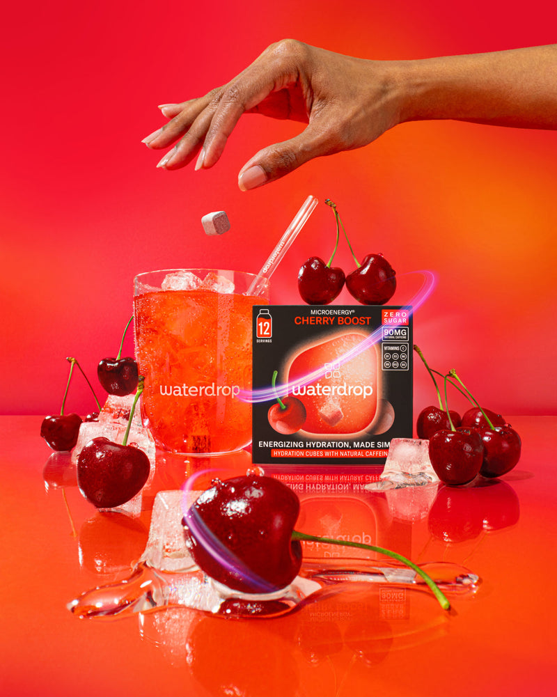 Energieboostset: Een hand laat een Cherry Boost MicroEnergy-tablet in een glas met ijs en kersendrank vallen, omringd door verse kersen en de verpakking met zero sugar en 90 mg natuurlijke cafeïne.