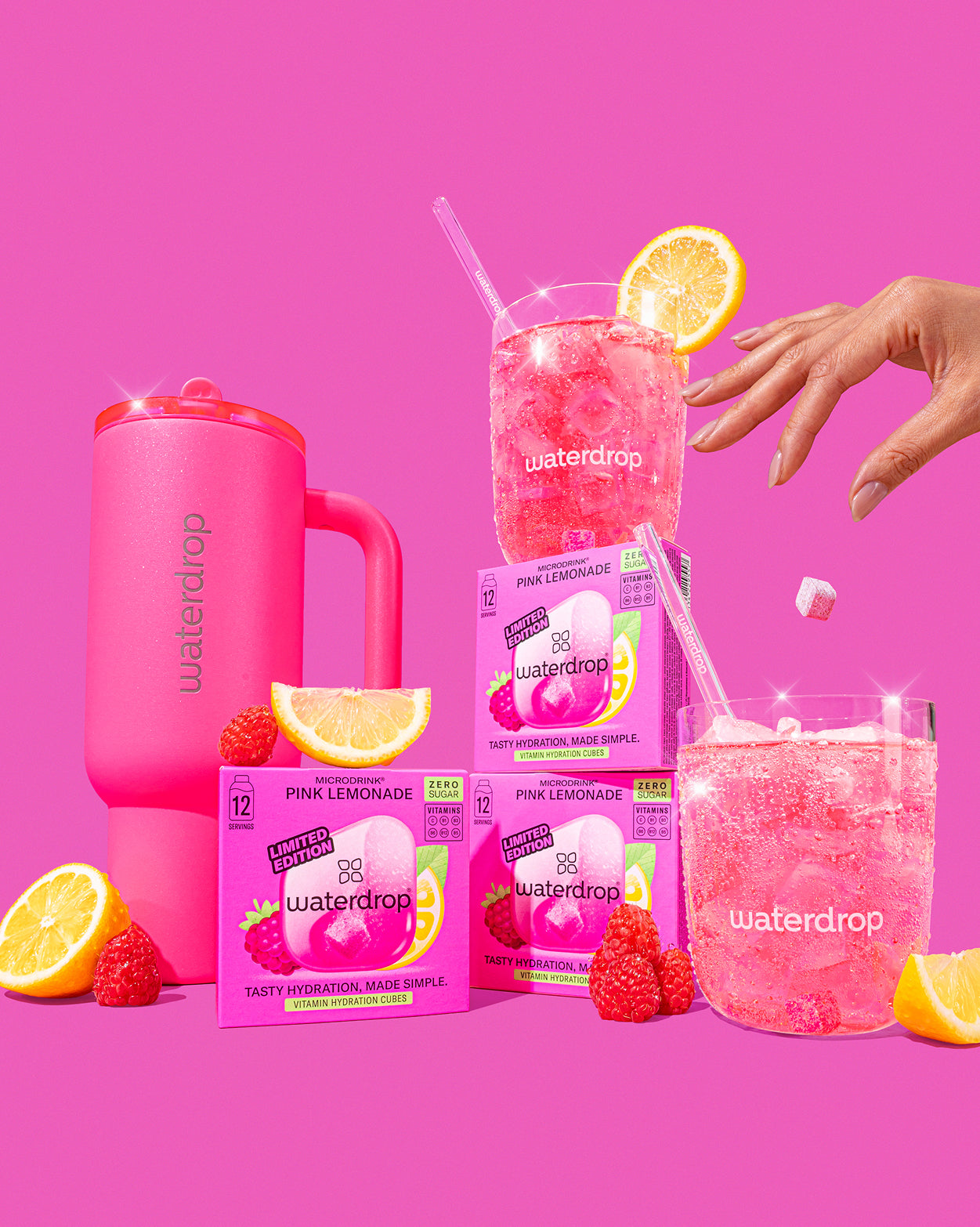 Naughty or Nice Hydration Set: Drie roze Waterdrop Pink Lemonade dozen, roze reismok, twee glazen met bruisend roze drankje en citroen, omringd door citroen- en frambozenpartjes. Hand voegt een cube toe.