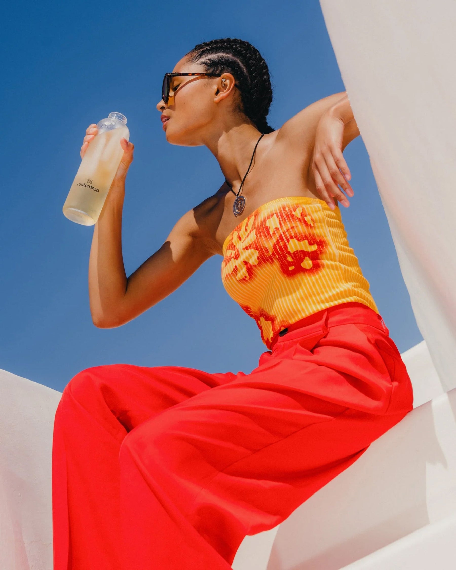 Een jong persoon drinkt uit een waterdrop fles, zittend onder een blauwe hemel. Ze dragen een strapless top en wijde broek, passend bij de Set: 3+1 Microdrinks fruitige ervaring.
