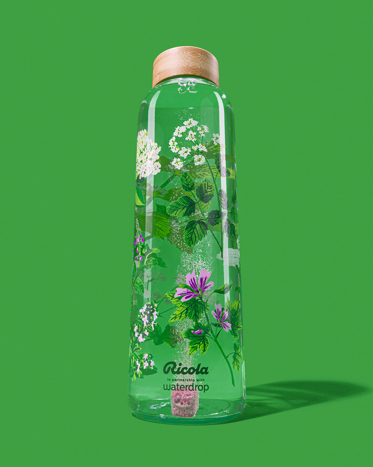 Hoge glazen fles met houten dop, bedrukt met Ricola's kruidenillustraties. Binnenin een bruisblokje dat oplost. Logo: Ricola in samenwerking met waterdrop. Ricola Edition Glass combineert frisheid en schoonheid.
