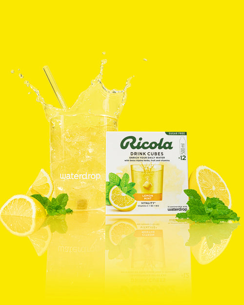 Een hoog glas citroen-muntwater met een waterdrop-herbruikbaar rietje, omringd door citroenpartjes en munt. Voor het glas staat een doosje Ricola Drink Cubes Citroen-Munt, suikervrij, met waterdrop-samenwerking.