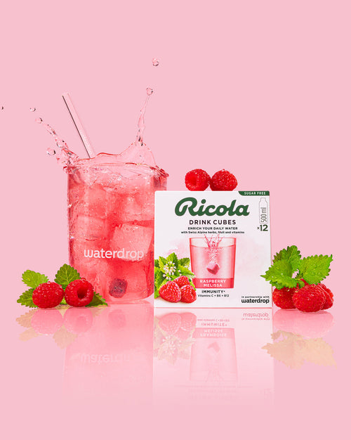 Exclusieve set waterdrop® x Ricola: Een helder glas met een roze frambozenmelisse drankje, ijs, en een splash; omringd door verse frambozen, melissablaadjes, en een doos Ricola Drink Cubes, suikervrij en immuniteitsversterkend.