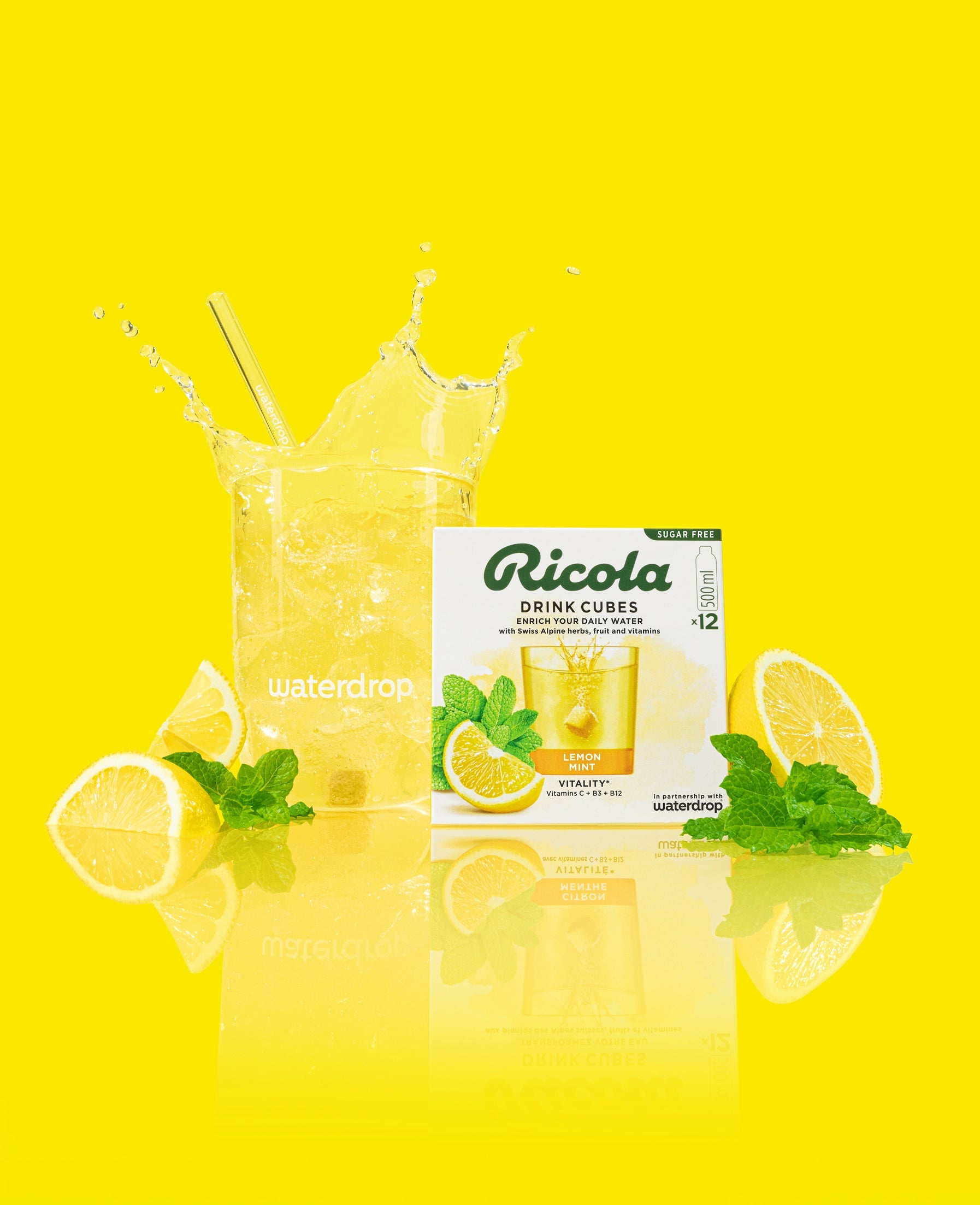 Exclusieve set waterdrop® x Ricola: Een helder waterdrop-glas gevuld met een gele drank, omringd door een Ricola Drink Cubes doos (Citroen Munt, suikervrij), citroenpartjes en munt, tegen een stralende gele achtergrond.