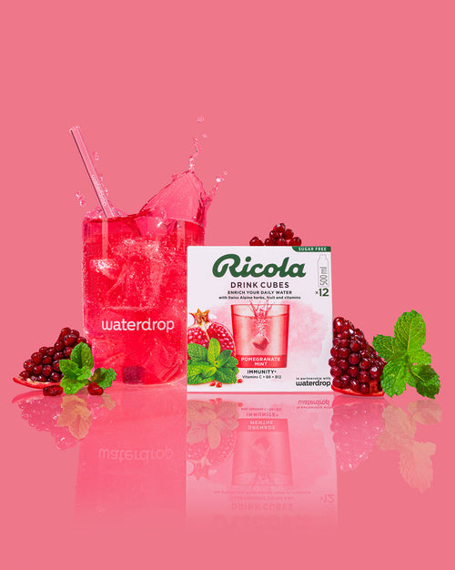 Granaatappel-Munt waterverbeteraar: een glas met roze/rode drank, ijsblokjes en een splash, omringd door granaatappelpitjes, verse munt en Ricola Drink Cubes, suiker- en calorievrij, vitamine C, B6, B12.