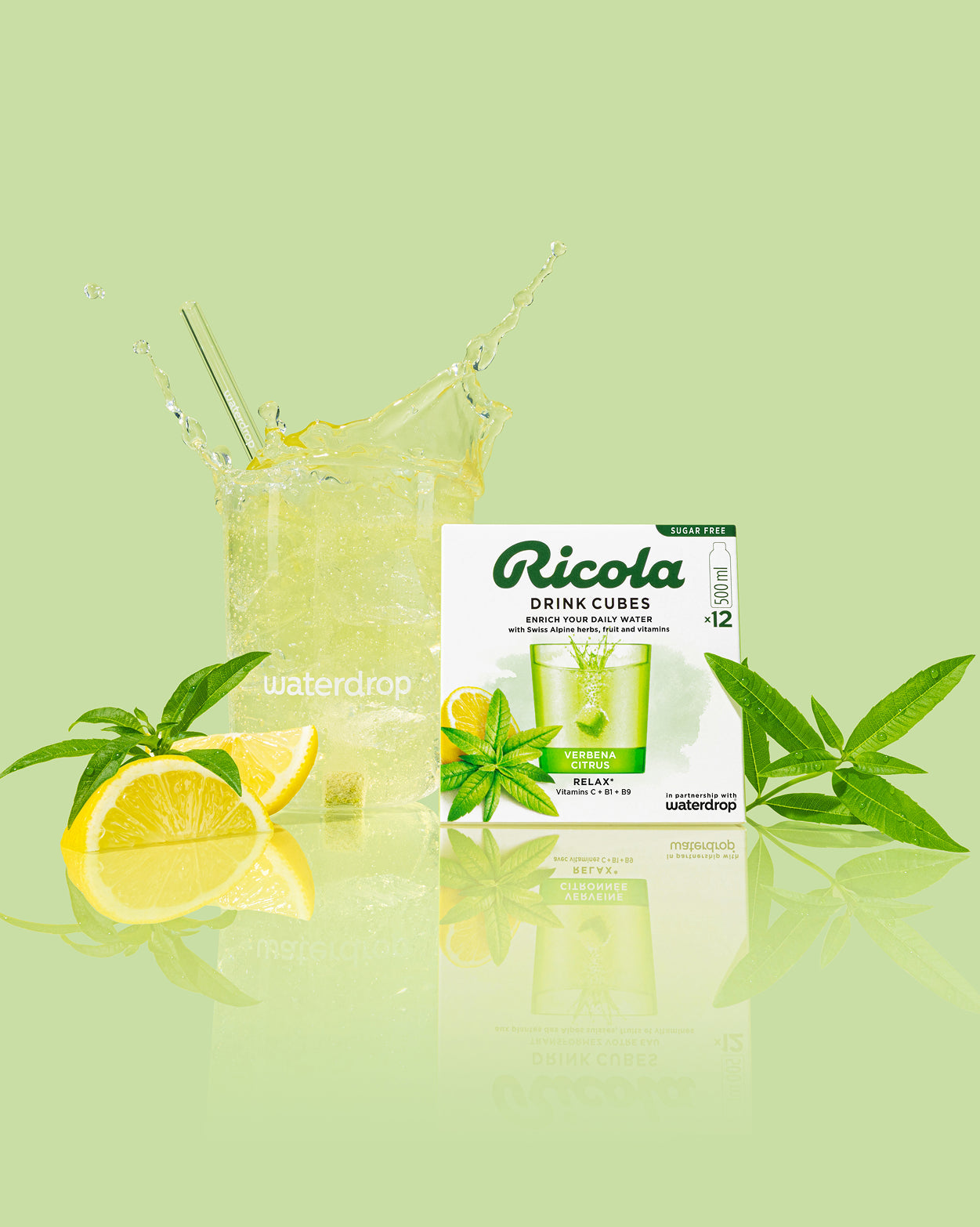 Een glas met ijskoude, geelgroene bruisende Verveine-Citrus water, met een waterdrop-rietje. Ernaast liggen citroenpartjes en verbena. Rechts staat een doos Ricola Drink Cubes Verbena Citrus smaak, suikervrij.