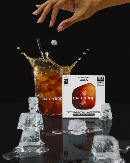 Prikkelend Trio Set: een close-up van een hand die een doos met de bruisende smaken SINAASAPPEL, COLA en FROSTED LIME vasthoudt, naast ijsblokjes op tafel, perfect voor bruiswater.