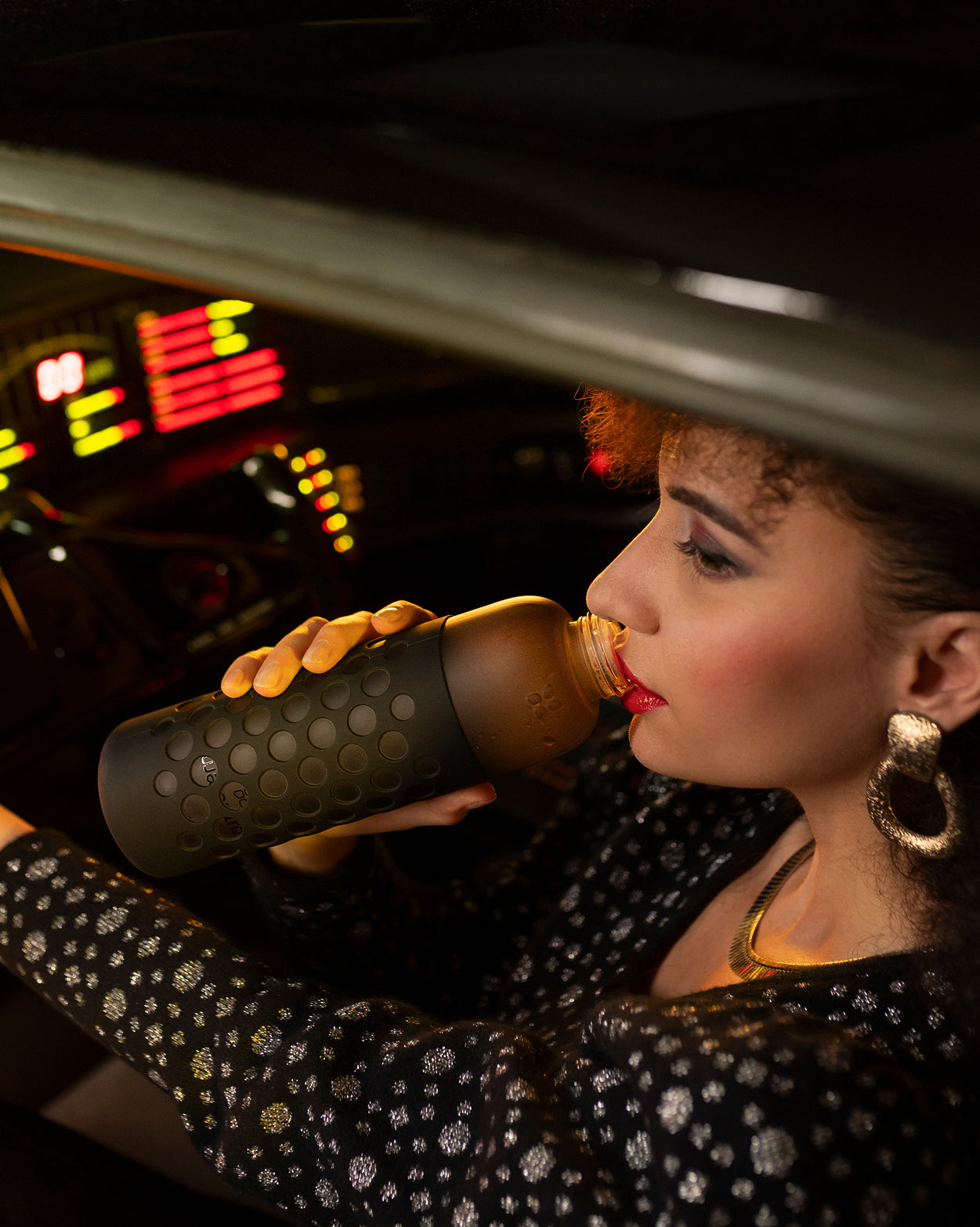 Vrouw in auto neemt een slok uit de Glazen set Booster, een slanke glazen fles met donkere siliconen hoes. Haar glamoureuze look en de sfeervolle verlichting creëren een retrofuturistische ambiance.