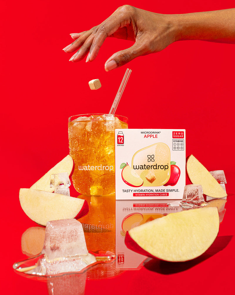 Limited Edition Twin Pack van Waterdrop: Een glas met ijs en een amberkleurige drank, met een hand die een waterdrop® Apple Microdrink hydratieblokje laat vallen, naast een verpakking van 12 porties.
