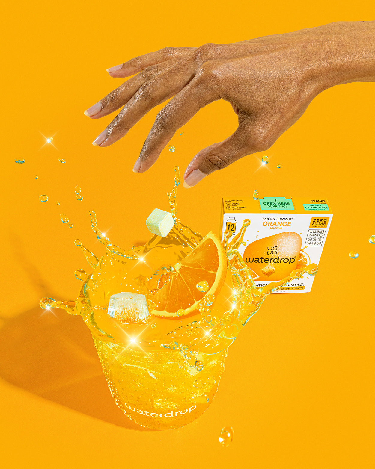 SINAASAPPEL 2+1 SET: Een hand boven een glas bruisend water voegt een waterdrop Microdrink tablet toe, met een sinaasappelschijf en spetters. Achtergrond toont de bijbehorende productdoos, ideaal voor verfrissende hydratatie.