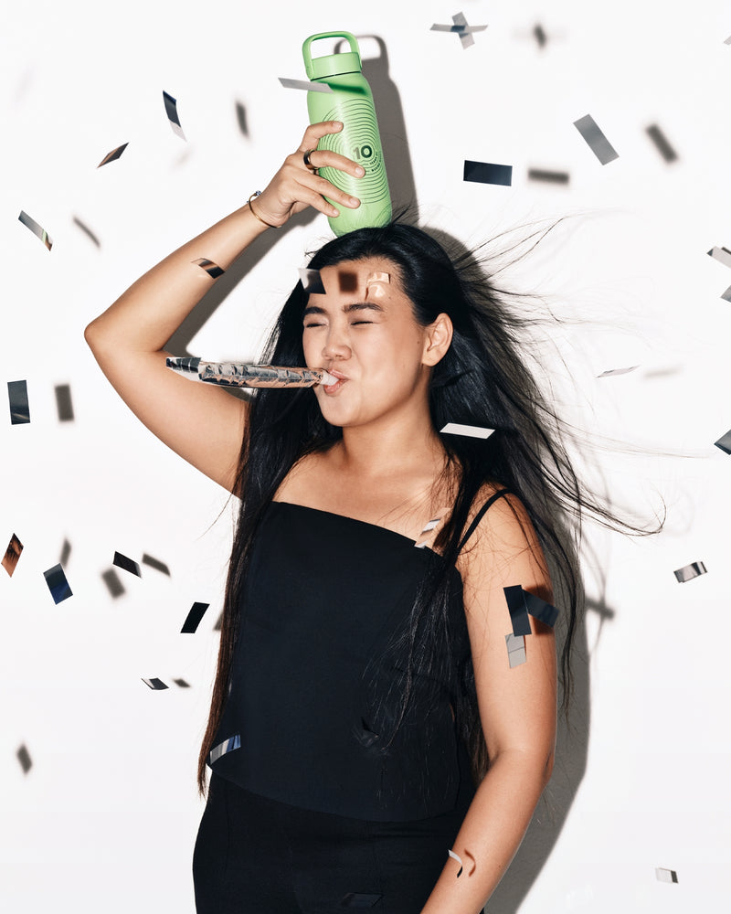 Vrouw in zwart draag een groene fles boven haar hoofd terwijl confetti naar beneden valt.