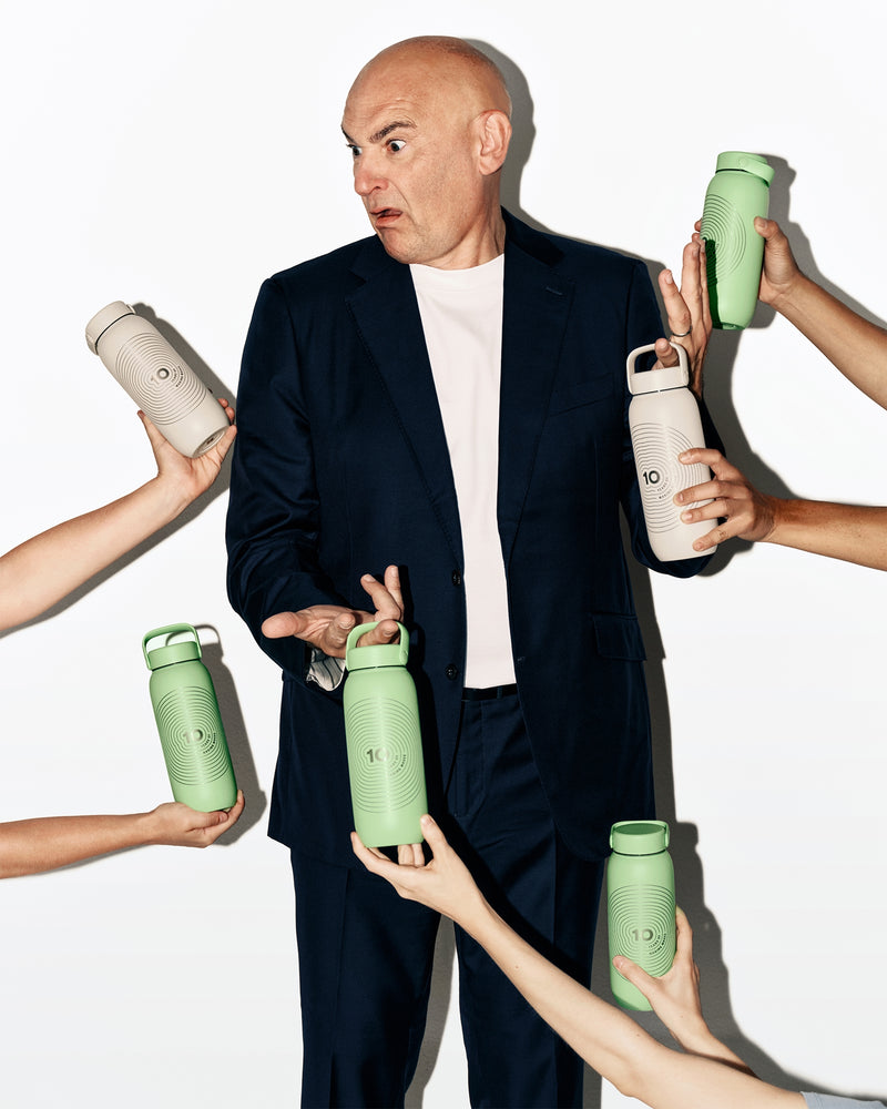 Een man in een donker pak staat terwijl meerdere handen groene en witte waterflessen om hem heen vasthouden.