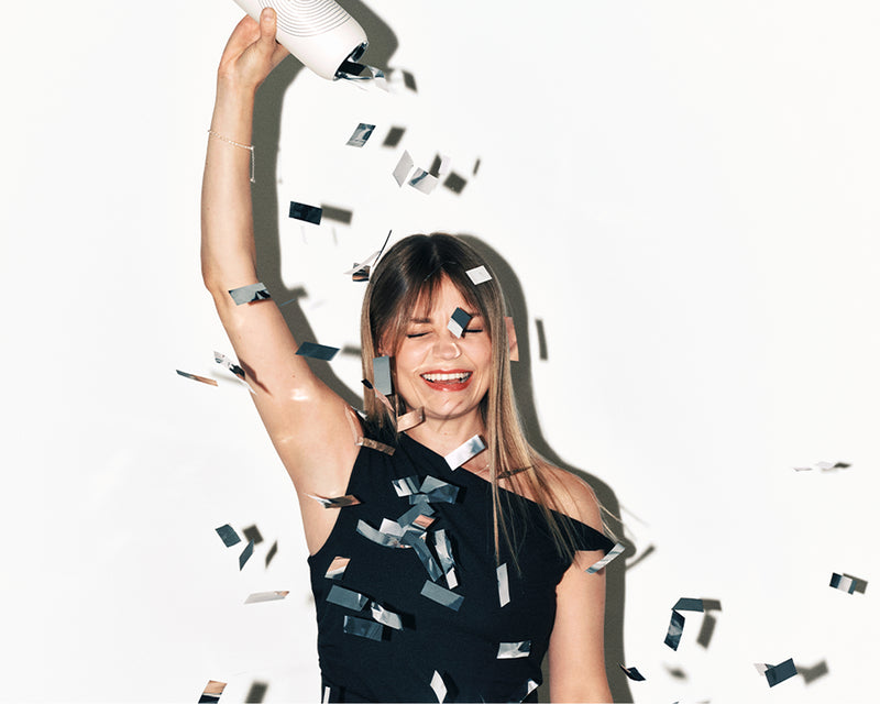 Vrouw in een zwarte off-shoulder jurk glimlacht terwijl ze confetti uit een party popper laat komen.