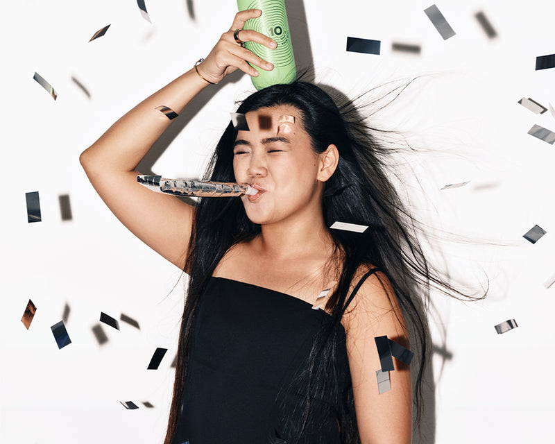 Vrouw in een zwarte jurk viert met een feesthoorn en een groene spuitbus, omringd door confetti.