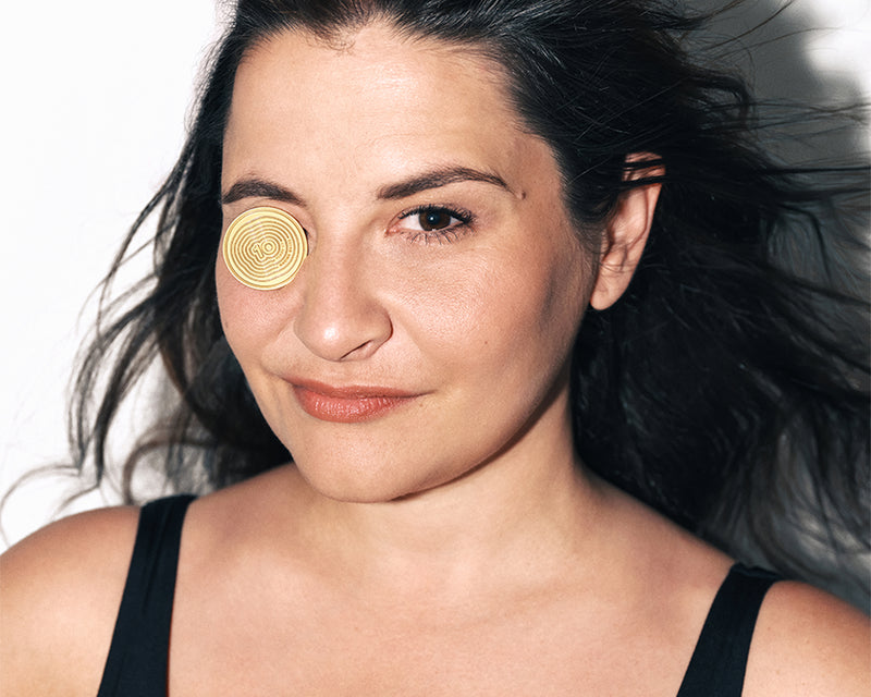 Vrouw met donker haar in een zwarte top, naar de camera gericht, met een gouden cirkelvormige patch op haar linkerwang bij het oog.