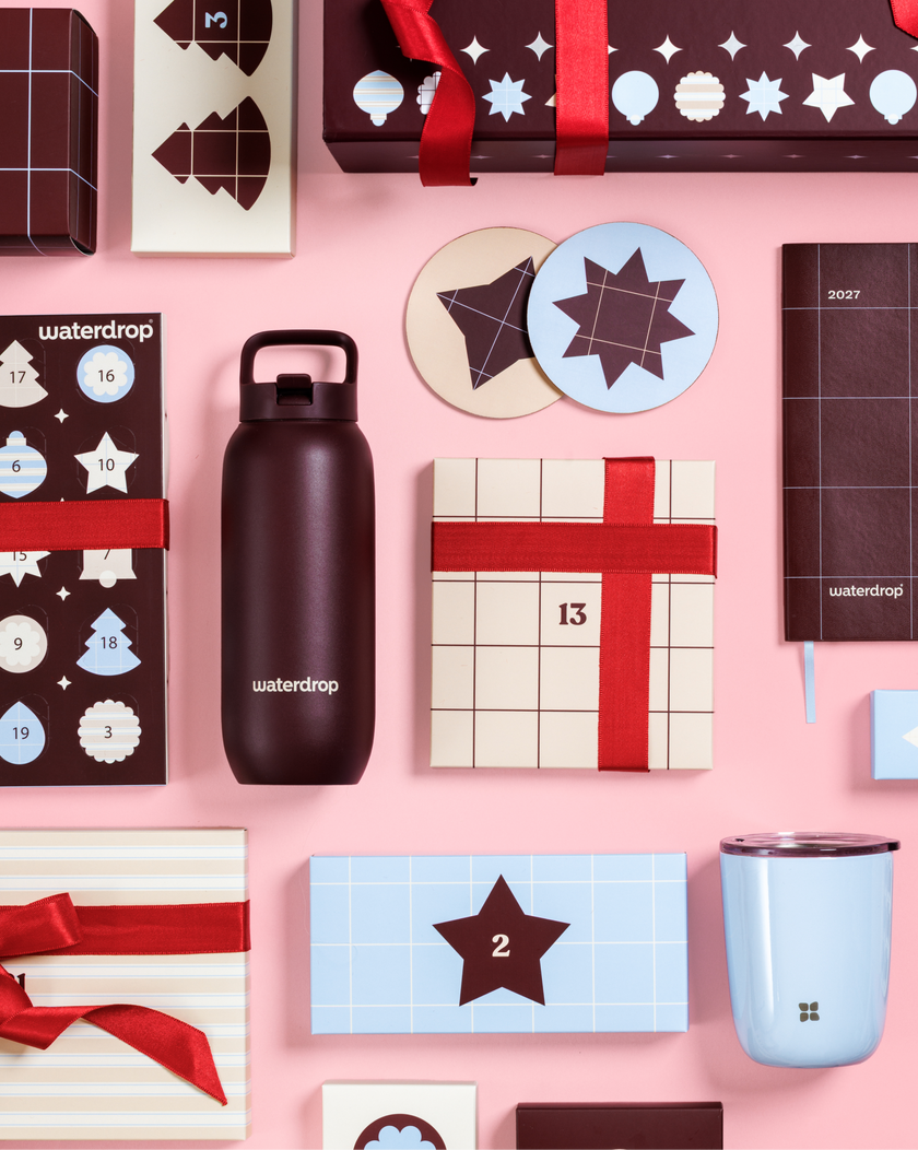 Diverse adventskalenders en cadeautjesdozen met rode linten op roze achtergrond, naast een zwarte waterfles en een lichtblauwe mok.