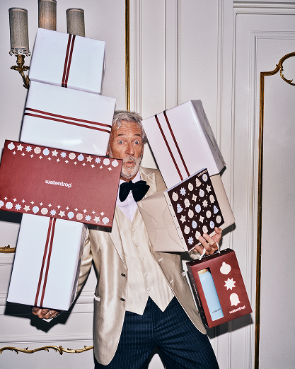 Man in een beige pak draagt meerdere ingepakte cadeaus binnen.