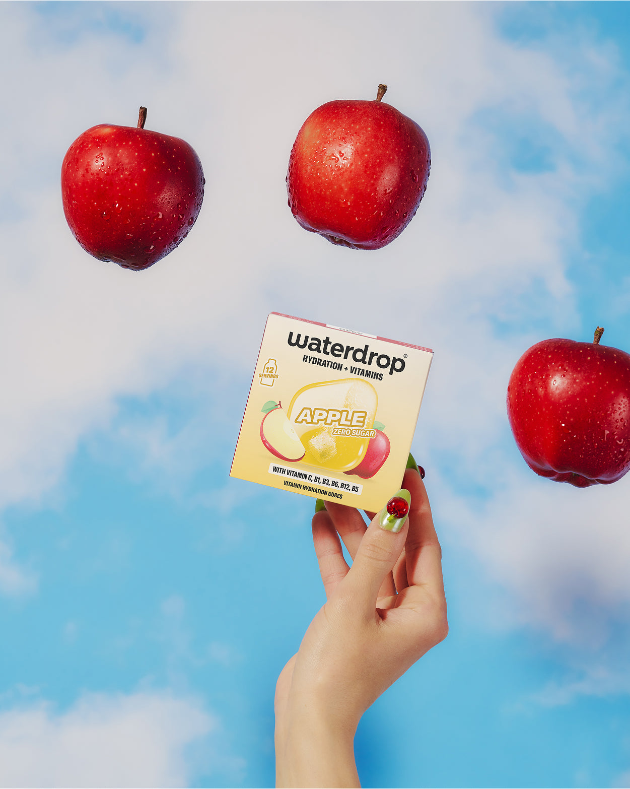 Hand houdt een kleine doos met een appelillustratie tegen een blauwe lucht met drie zwevende rode appels.