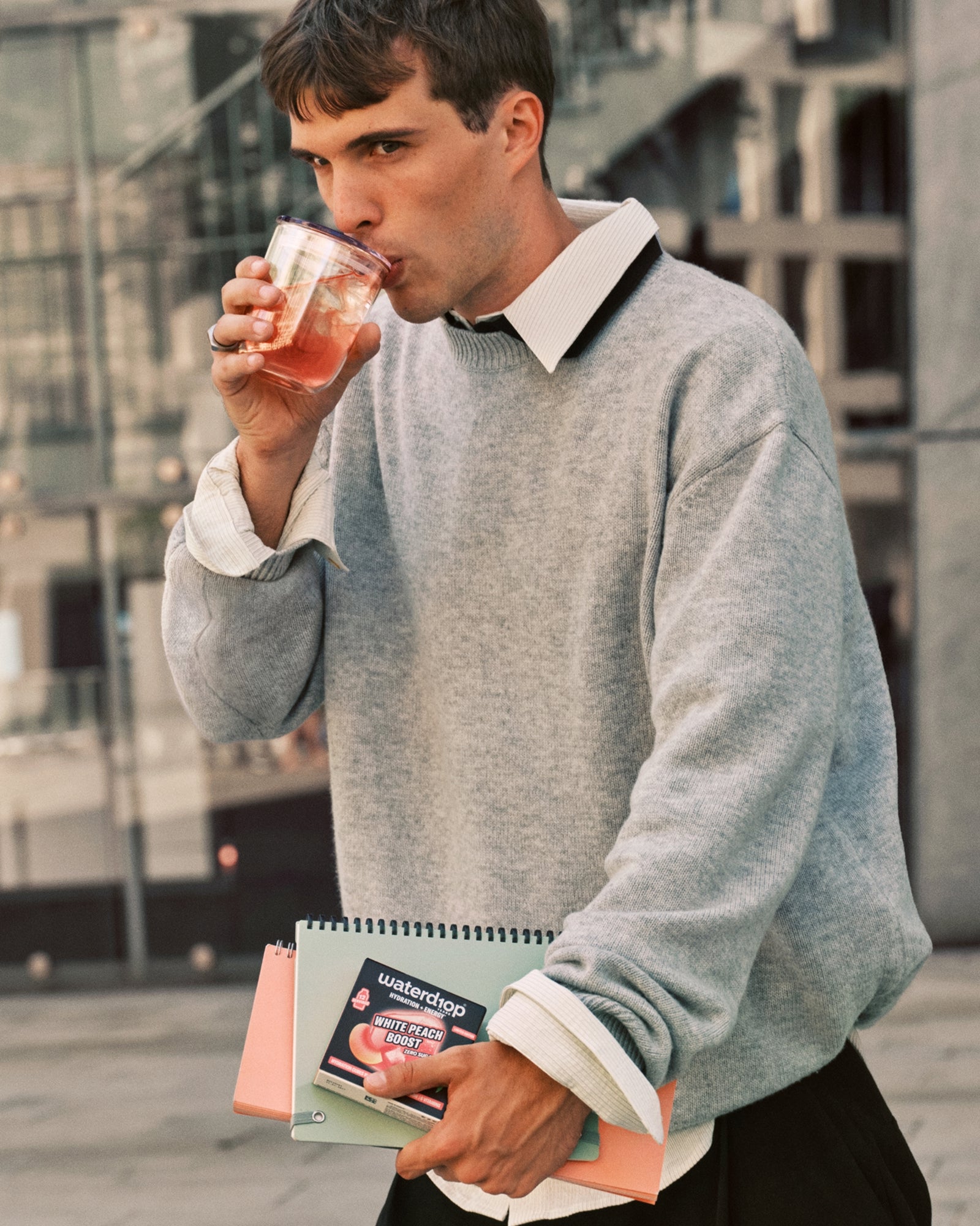 Man in grijze trui drinkt uit een beker terwijl hij notitieboeken draagt.