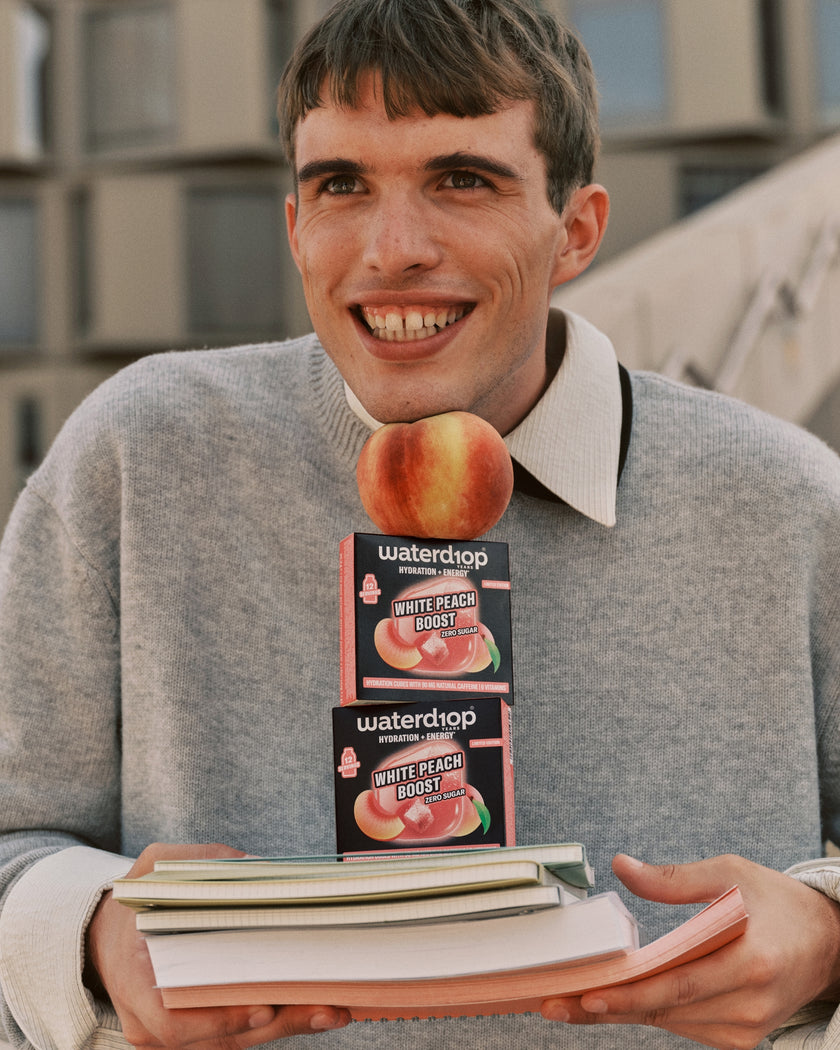 Jonge man in grijze trui houdt twee doosjes perzikensmaak boven een stapel boeken.