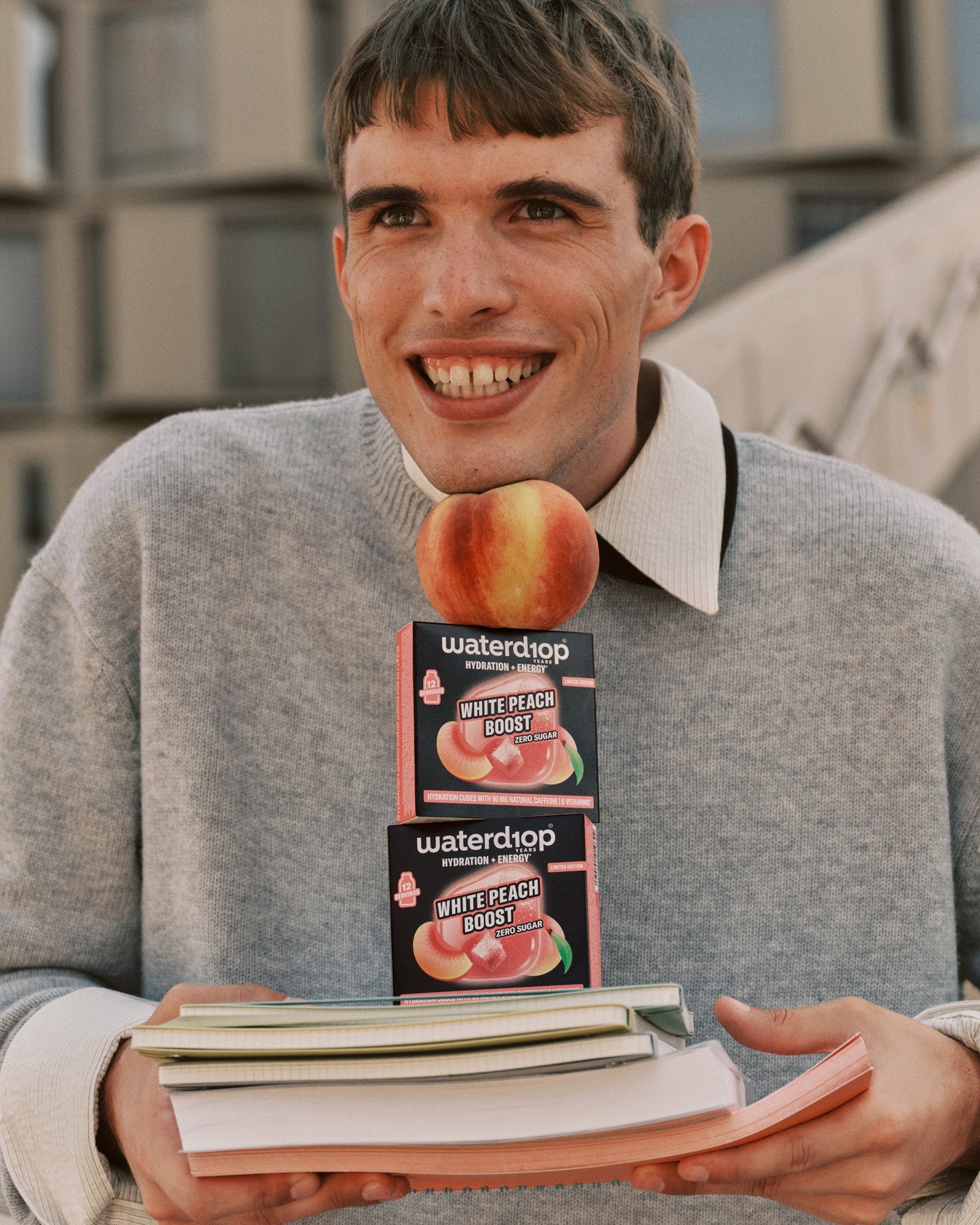 Jonge man in grijze trui houdt twee doosjes perzikensmaak boven een stapel boeken.