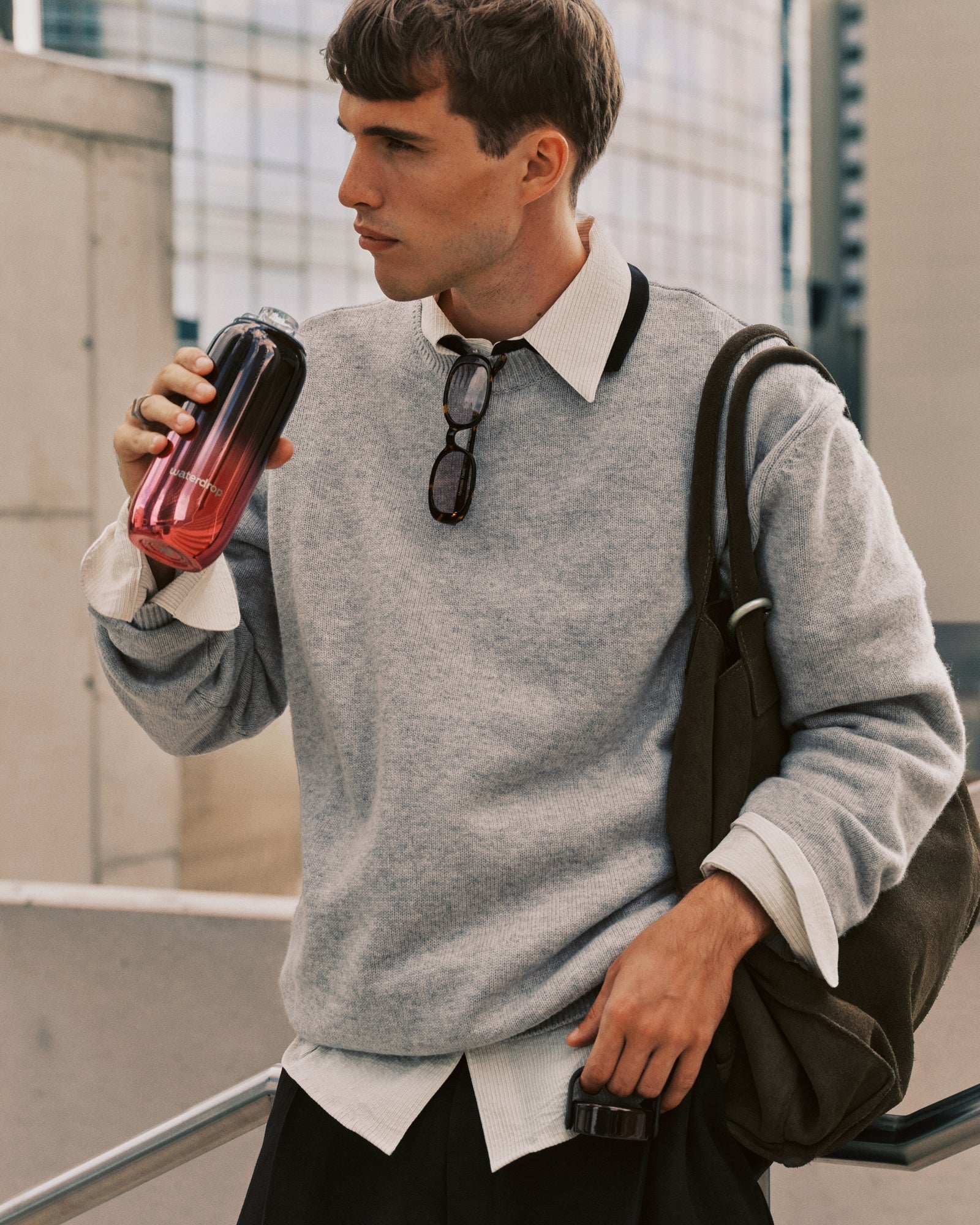 Jonge man in grijs trainingspak en wit overhemd met rugzak drinkt uit een glas met een kleurverloop.
