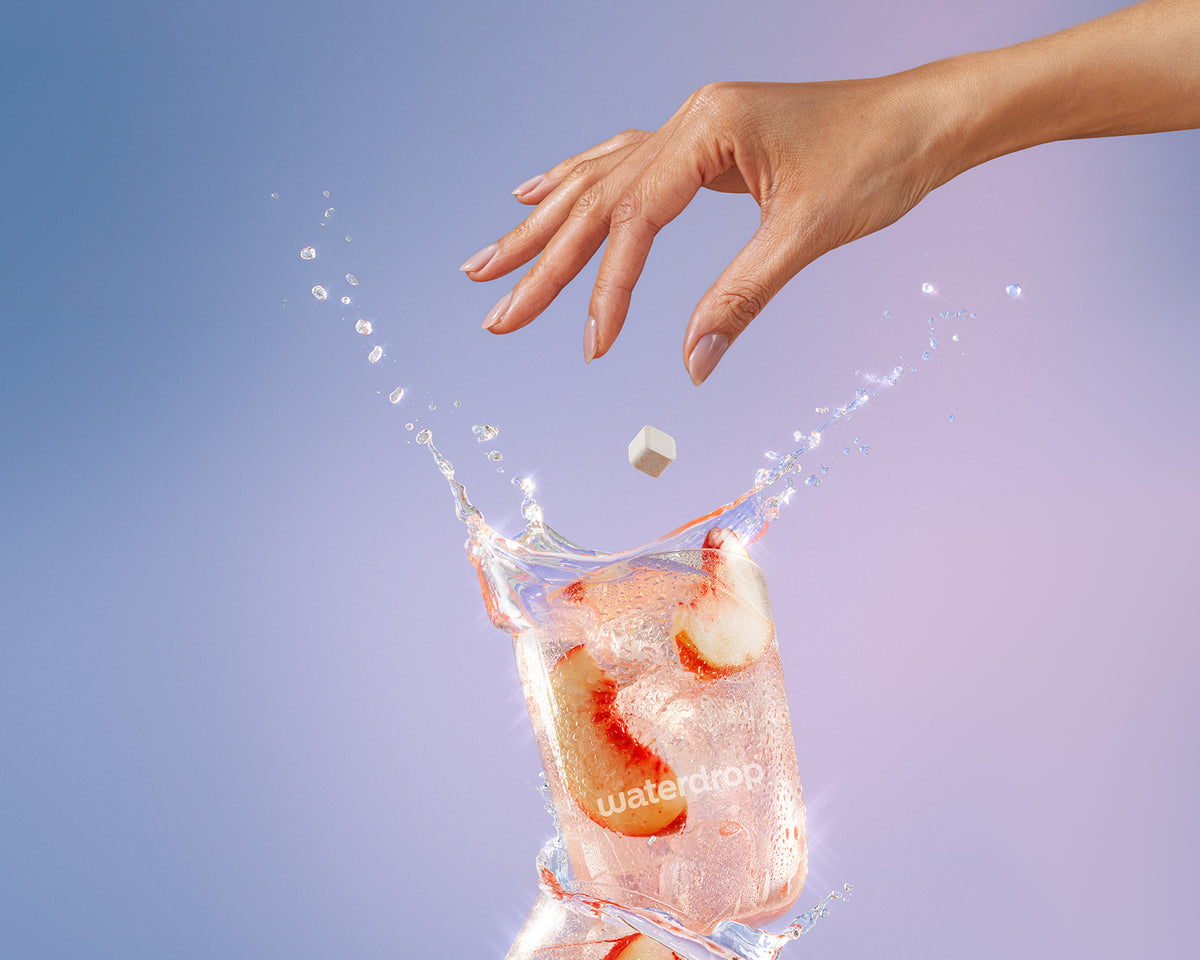 Een hand laat een kleine ijsblokje in een glas water met ijs vallen, waardoor het spat.